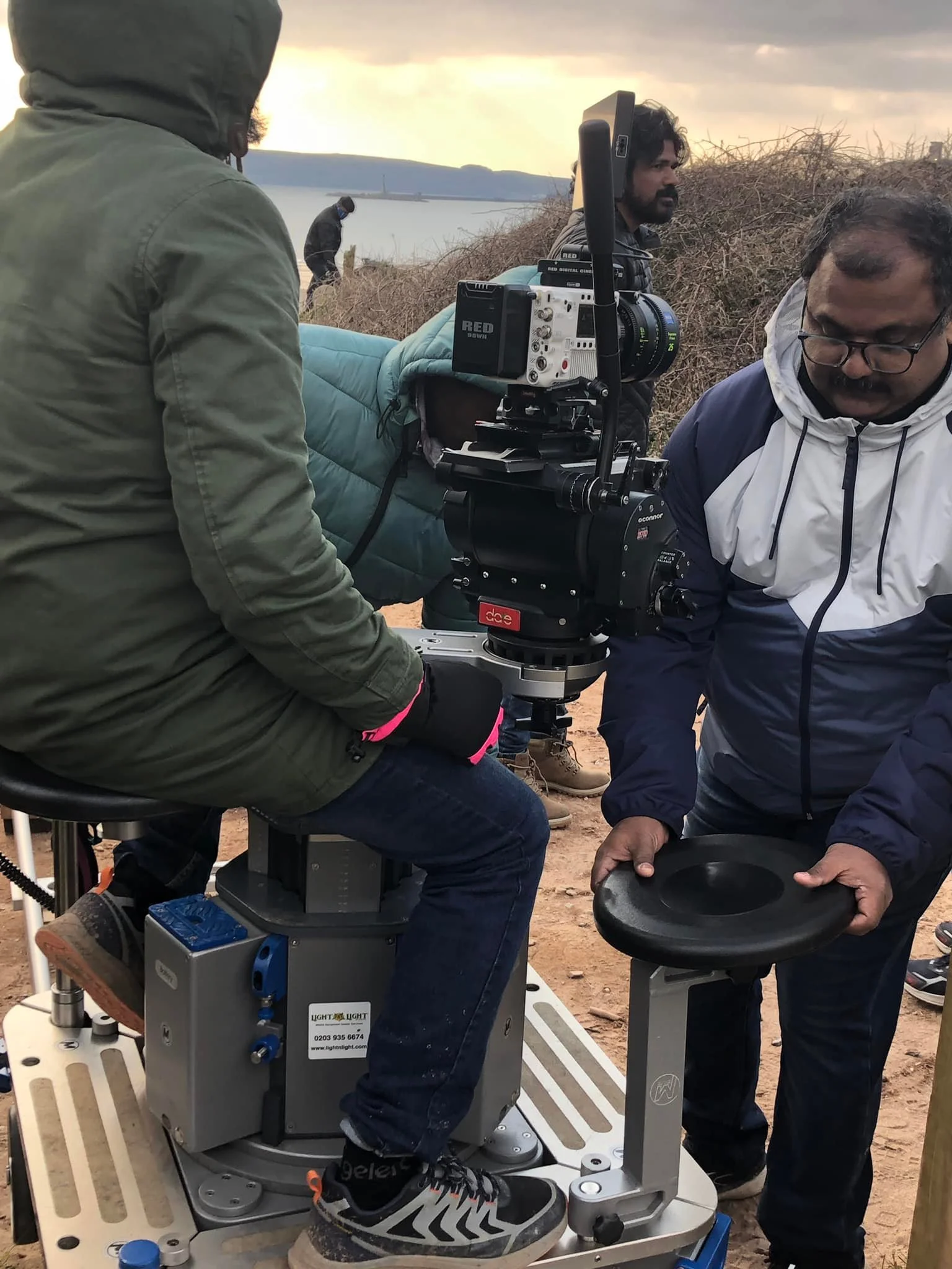 People working with a camera setup outdoors near a body of water during sunset, with one person seated on a chair and others assisting with equipment.