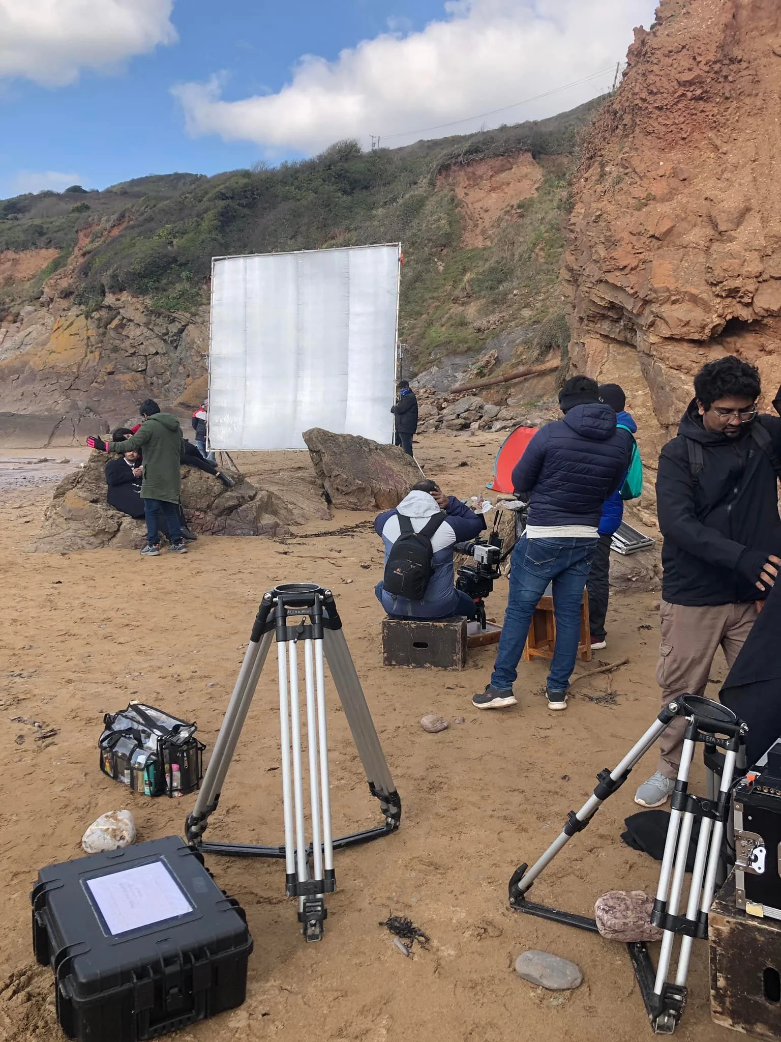 Film crew setting up equipment on a beach with rocky cliffs in the background, preparing for a shoot.