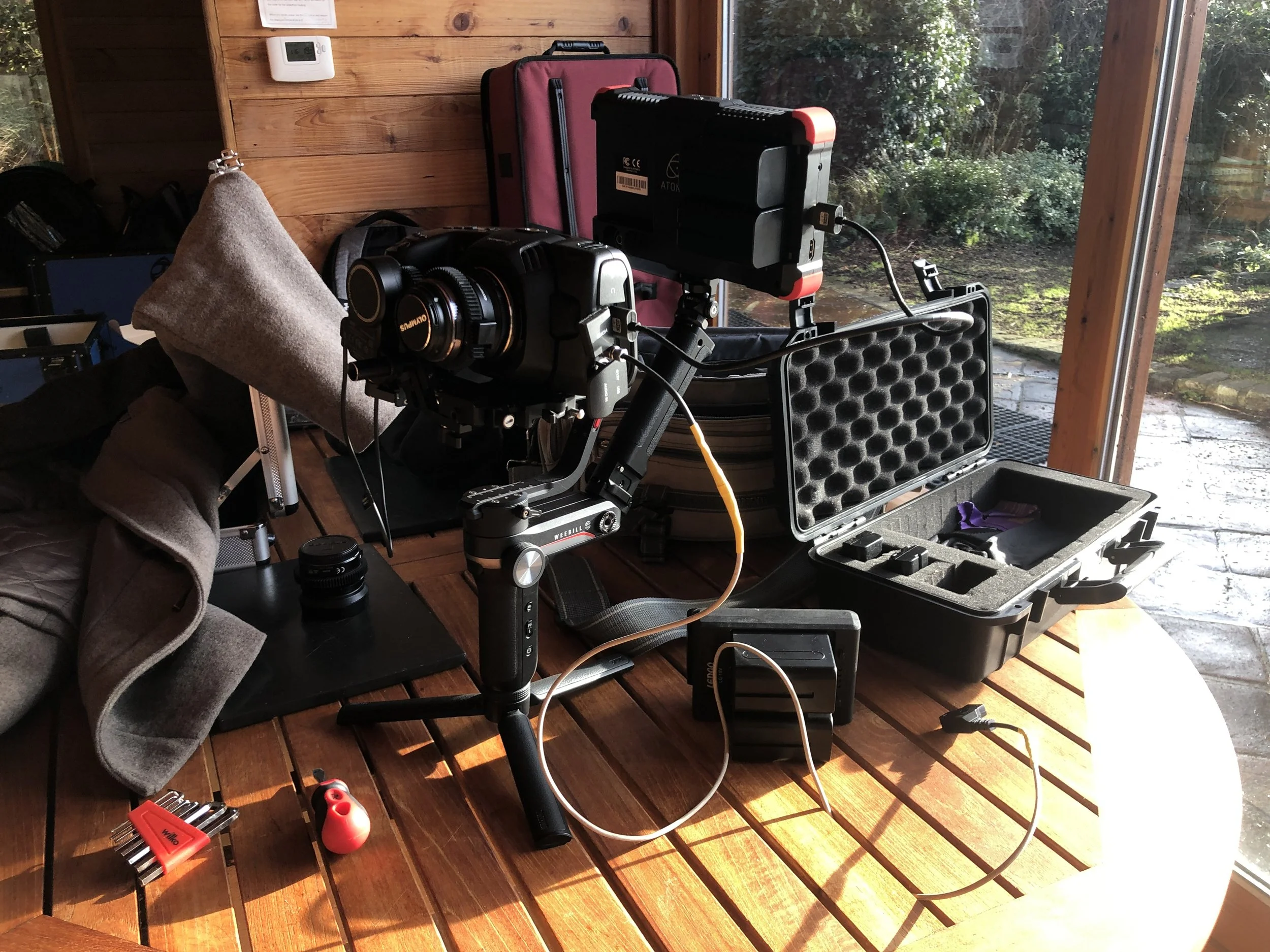 A camera mounted on a stabilizer rig with various camera equipment, a black camera case, and accessories on a wooden table near a large window with outdoor greenery visible.