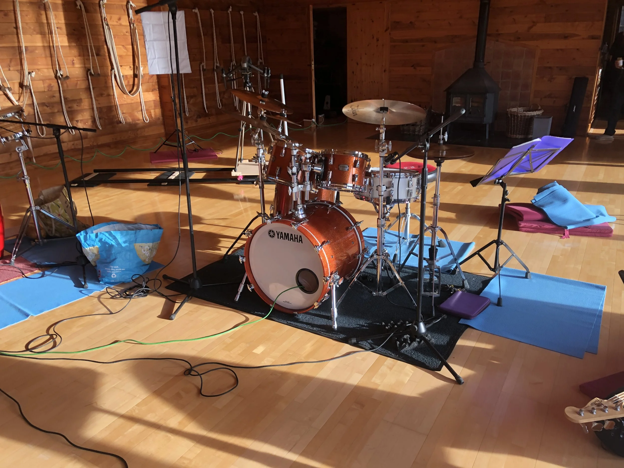 Drum set on a black rug surrounded by yoga mats and music stands in a wooden room.