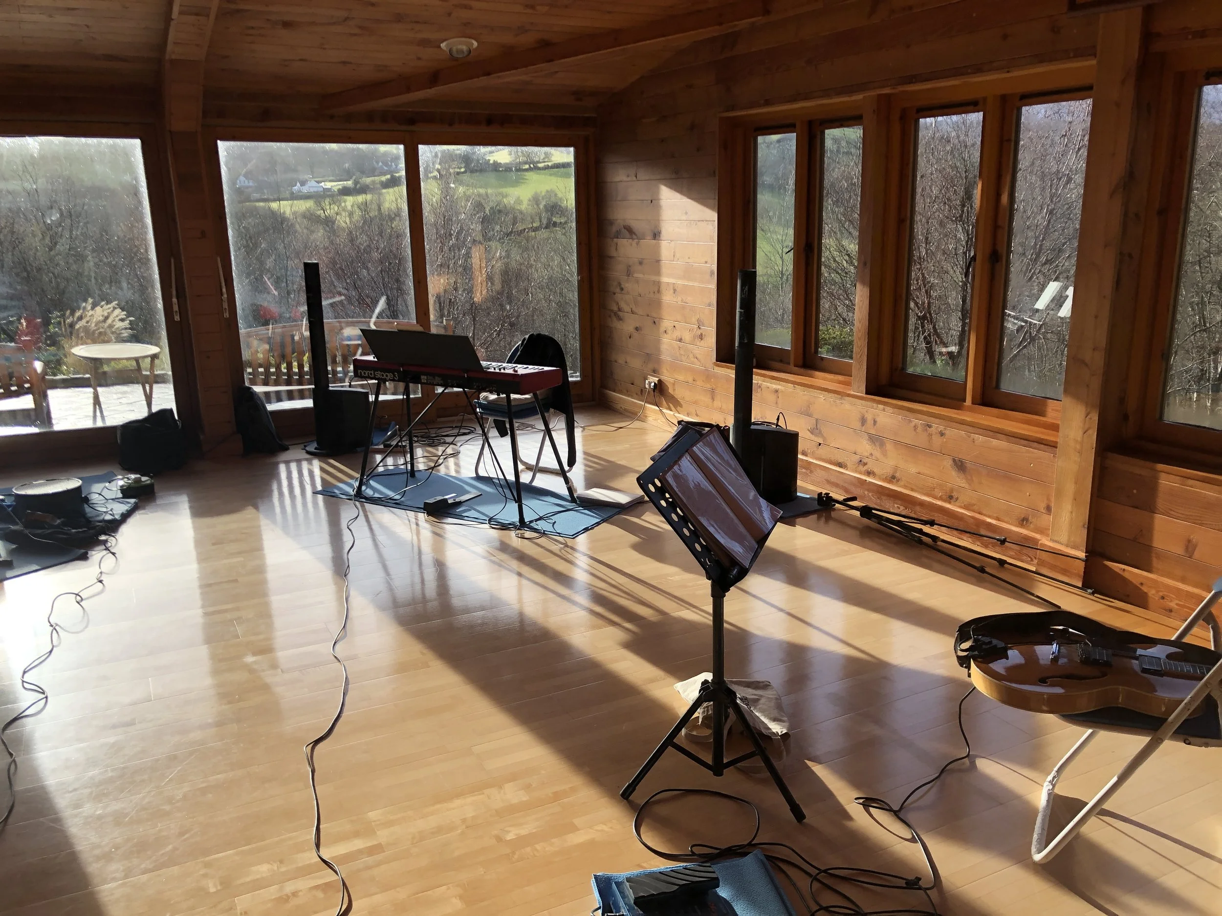 Empty wooden room with large windows, music stand, keyboard, guitar, and sound equipment, suggesting a music rehearsal or recording space.