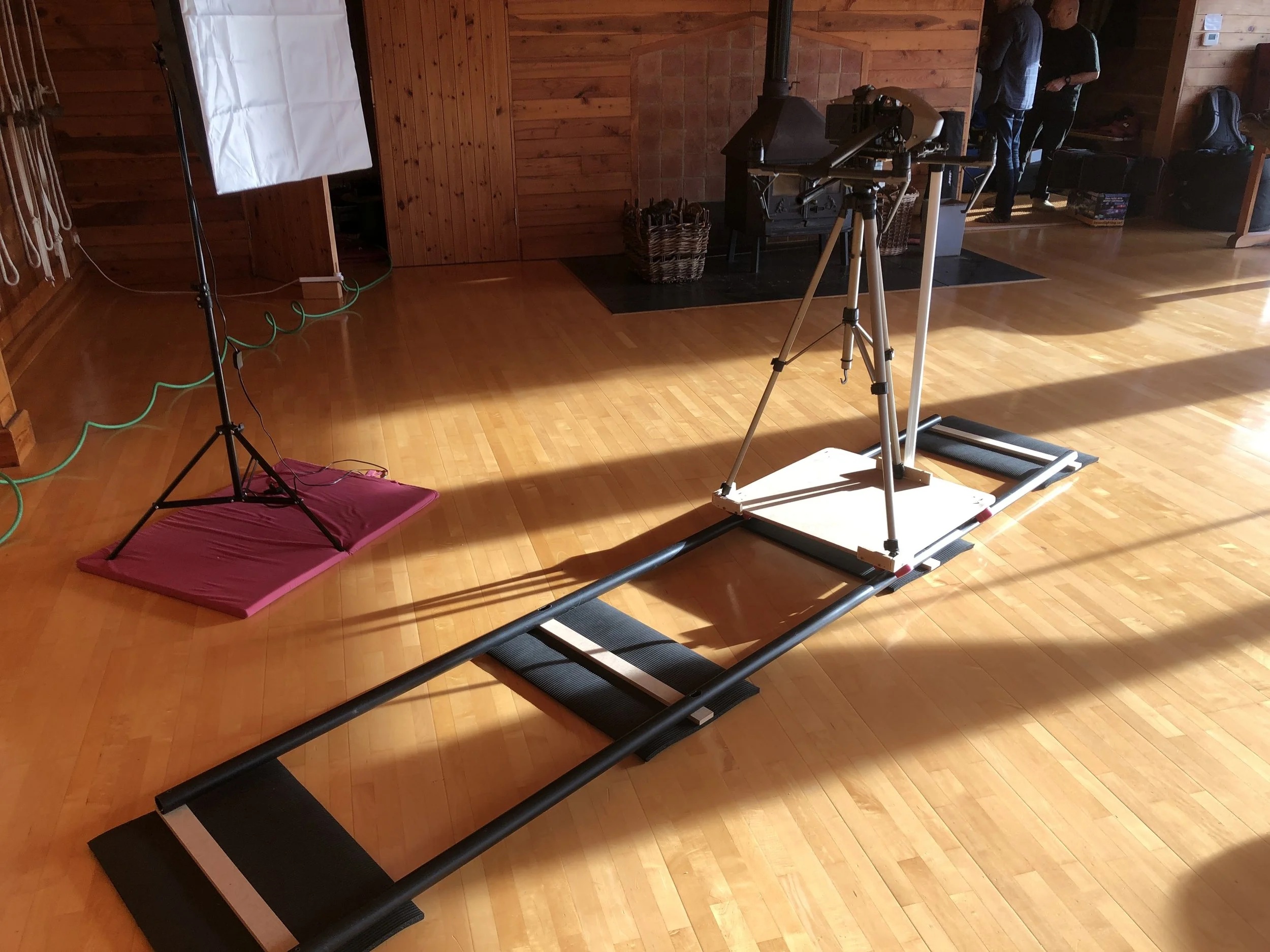 A treadmill with a camera mounted on it, set up in a room with wooden walls and floor, with photography equipment and people in the background.