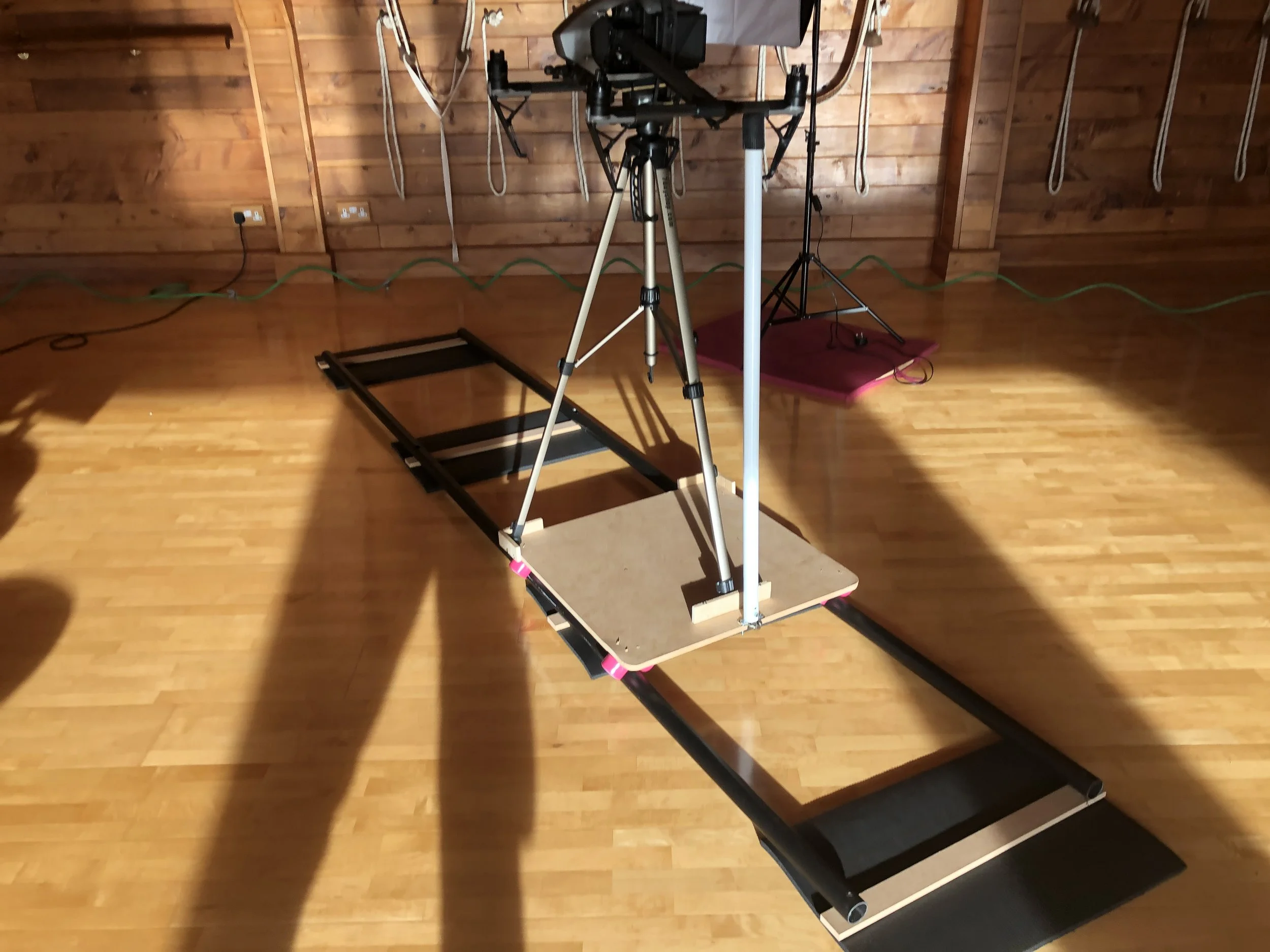 A treadmill with a camera system mounted on top, positioned on a rolling cart with a wooden platform, in a room with wooden walls and hardwood flooring, with green extension cords along the wall and photography equipment in the background.