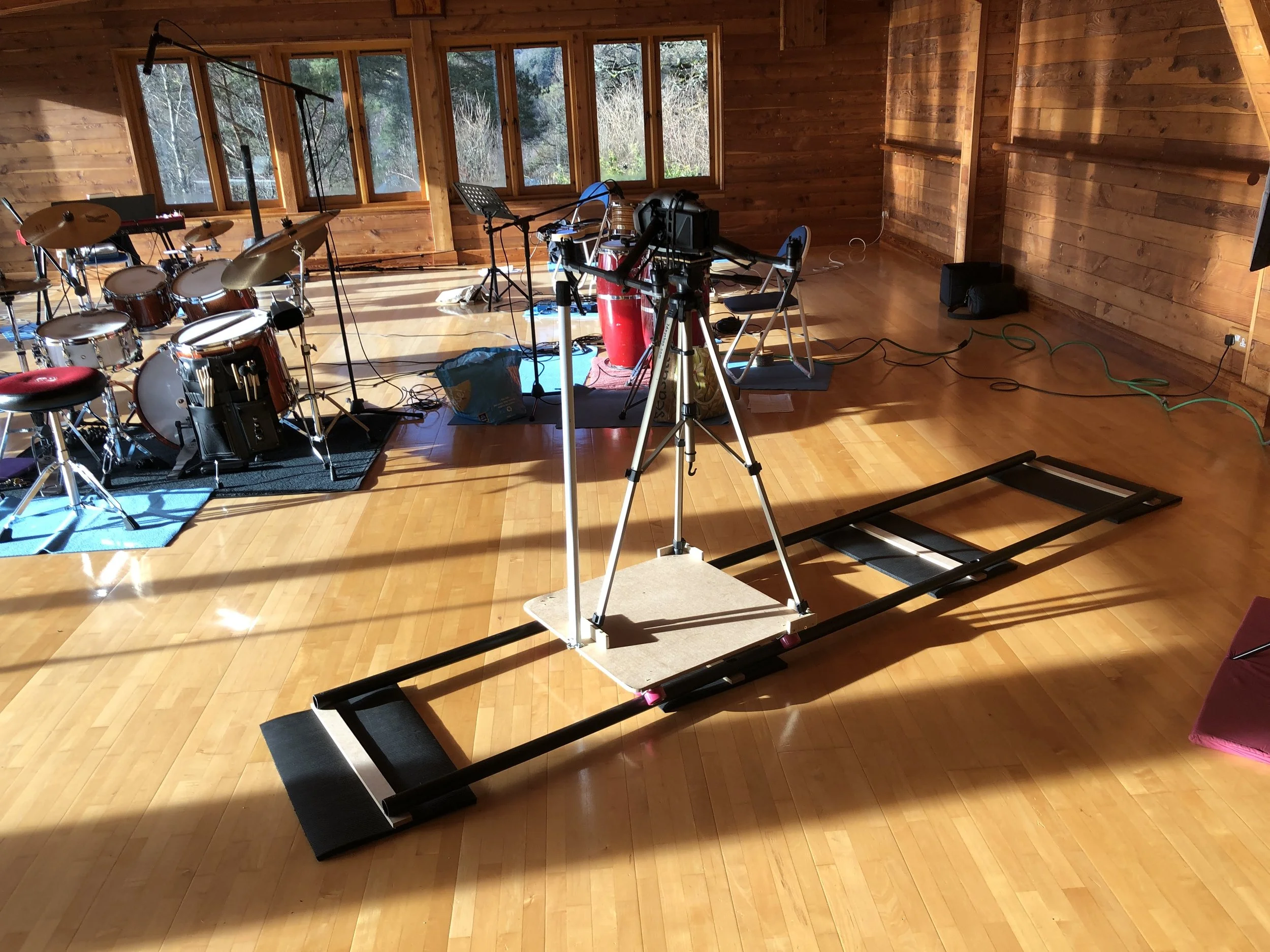 A music recording studio setup with drums on the left side and a treadmill-like device in the center for camera or equipment movement, with wooden walls and large windows in the background.