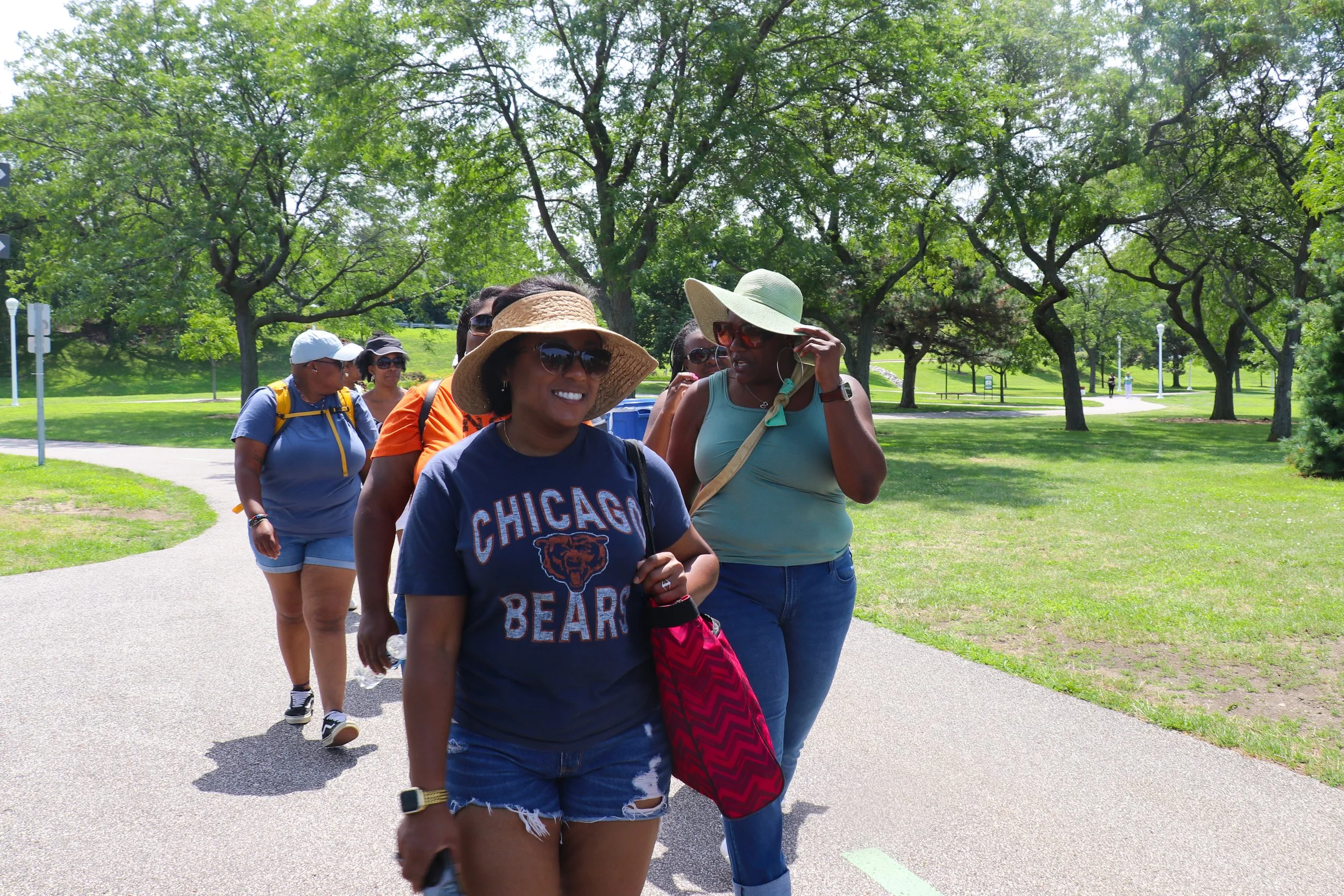A group of people walking in a park on a sunny day, wearing summer clothes and hats, with trees and green grass in the background.