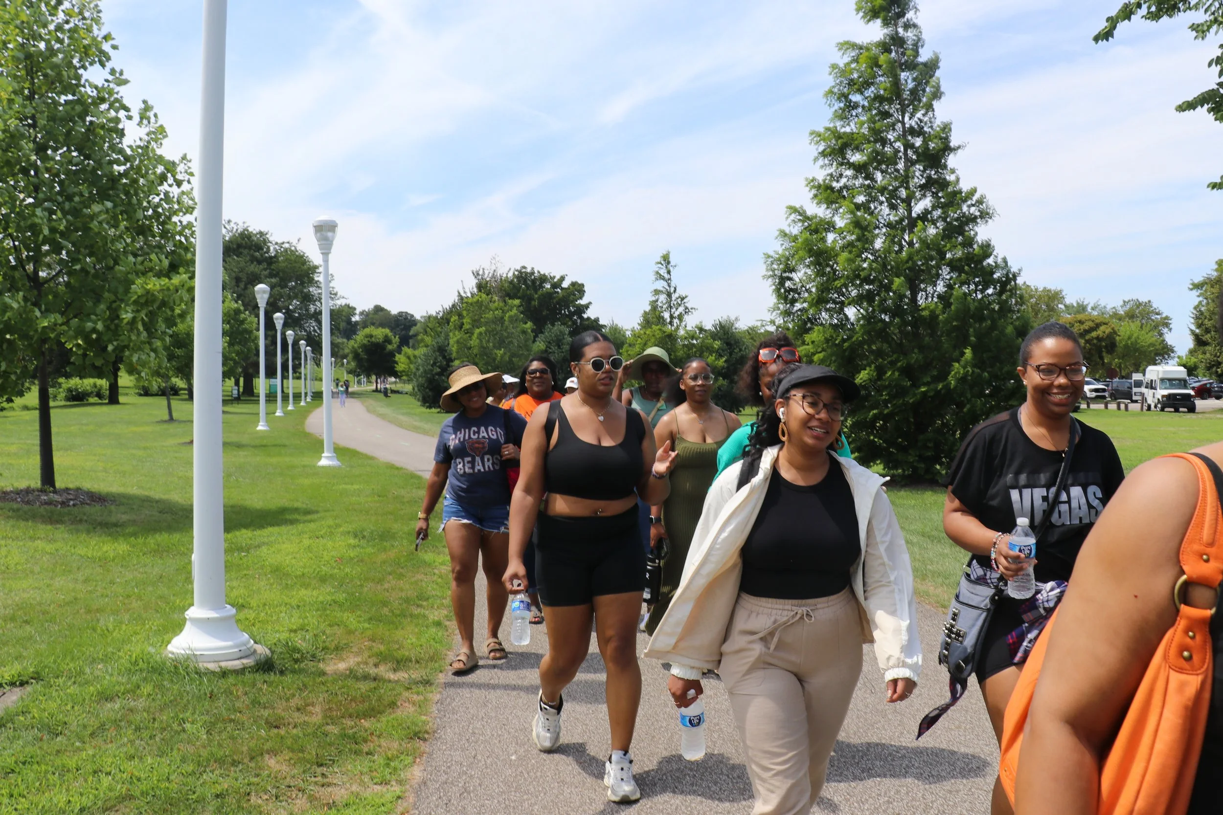 A group of women walking on a park trail on a sunny day, surrounded by green trees and grass.