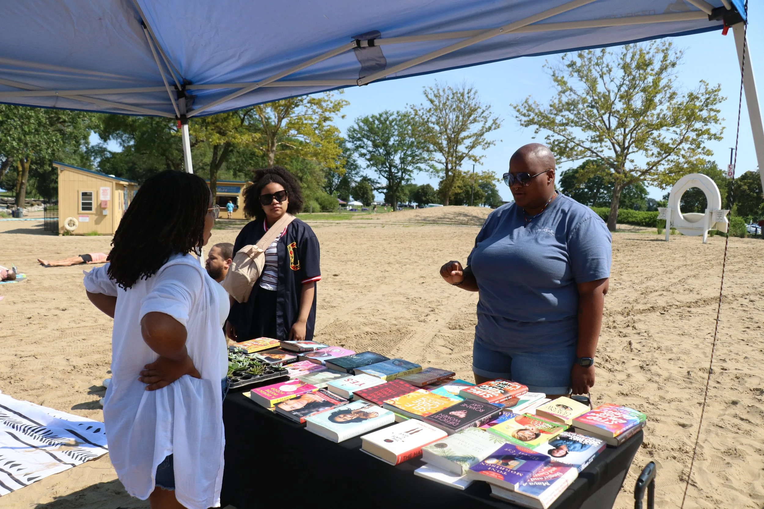 A woman at a book display table outdoors, talking to another woman at a book booth under a canopy, with trees and beach park in the background.