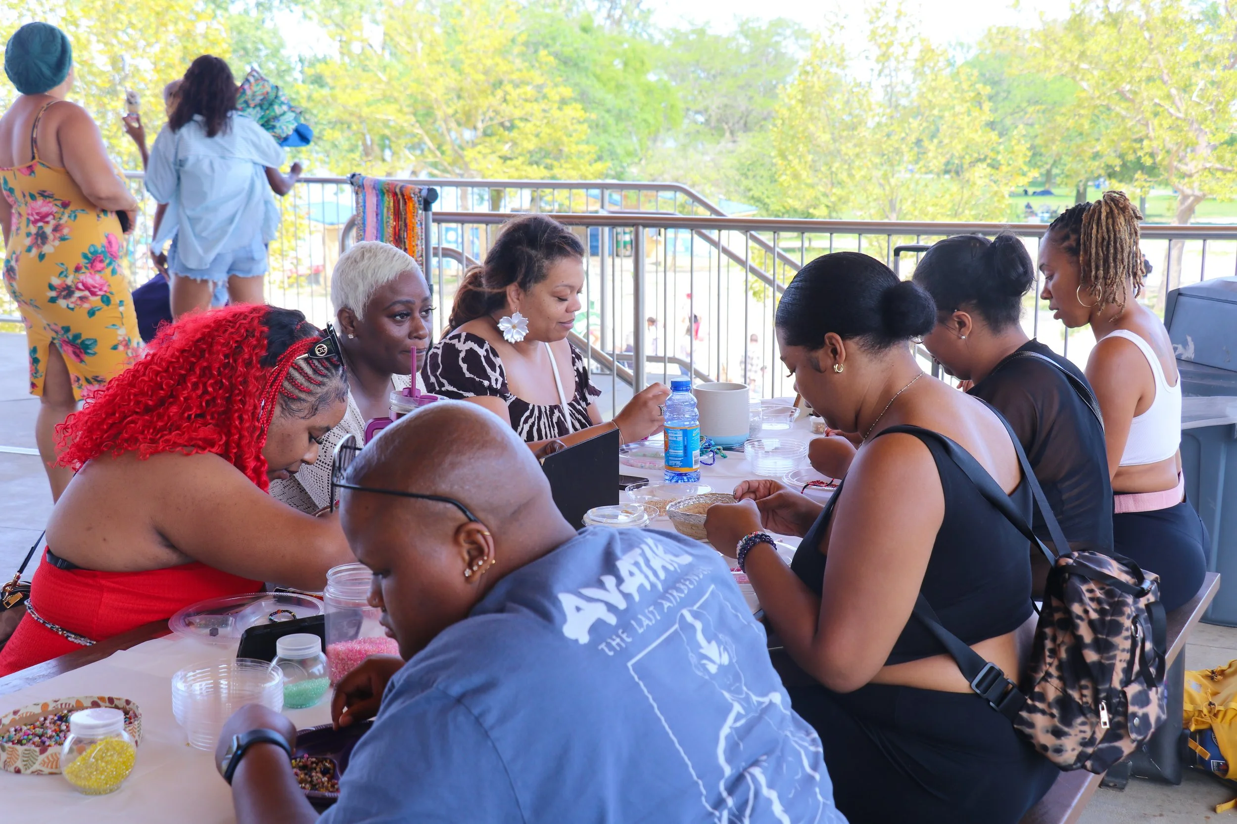 Group of women sitting at a table crafting jewelry outdoors on a bright, sunny day with trees in the background.