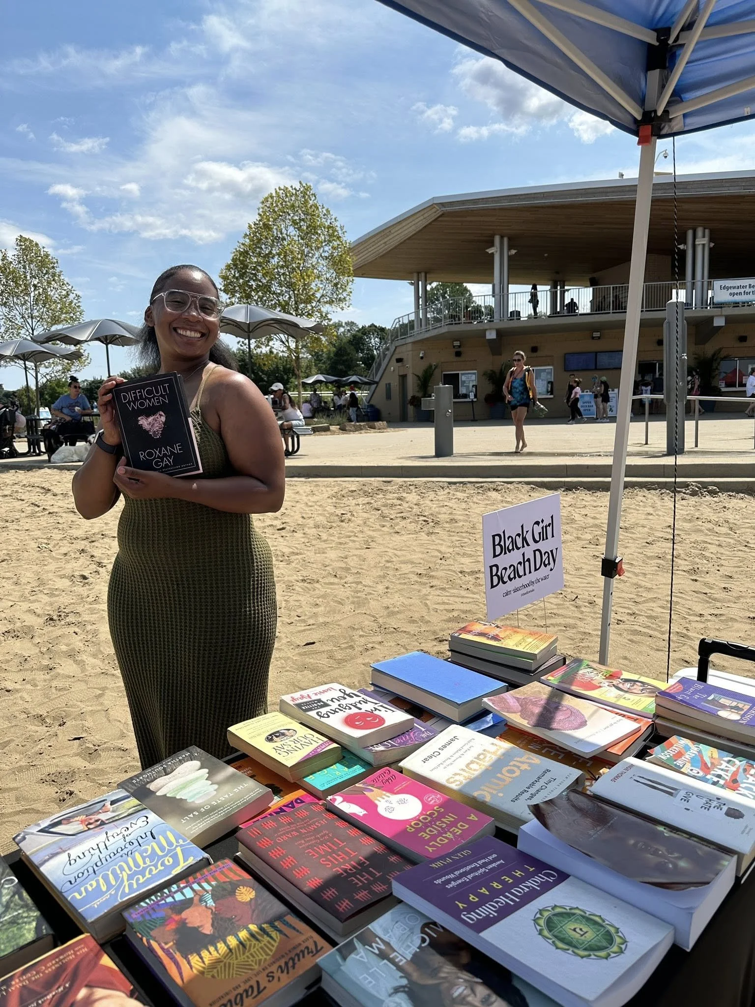 A smiling woman holding a book titled 'Difficult Women' standing next to a table with books at a Black Girl Beach Day event on a sandy beach with a building and people in the background.