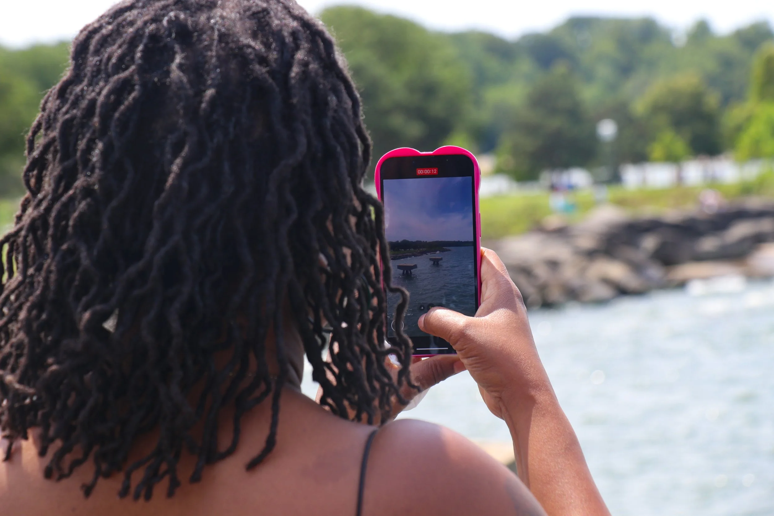 Person with dreadlocks taking a photo of a body of water with a smartphone at a scenic outdoor location.