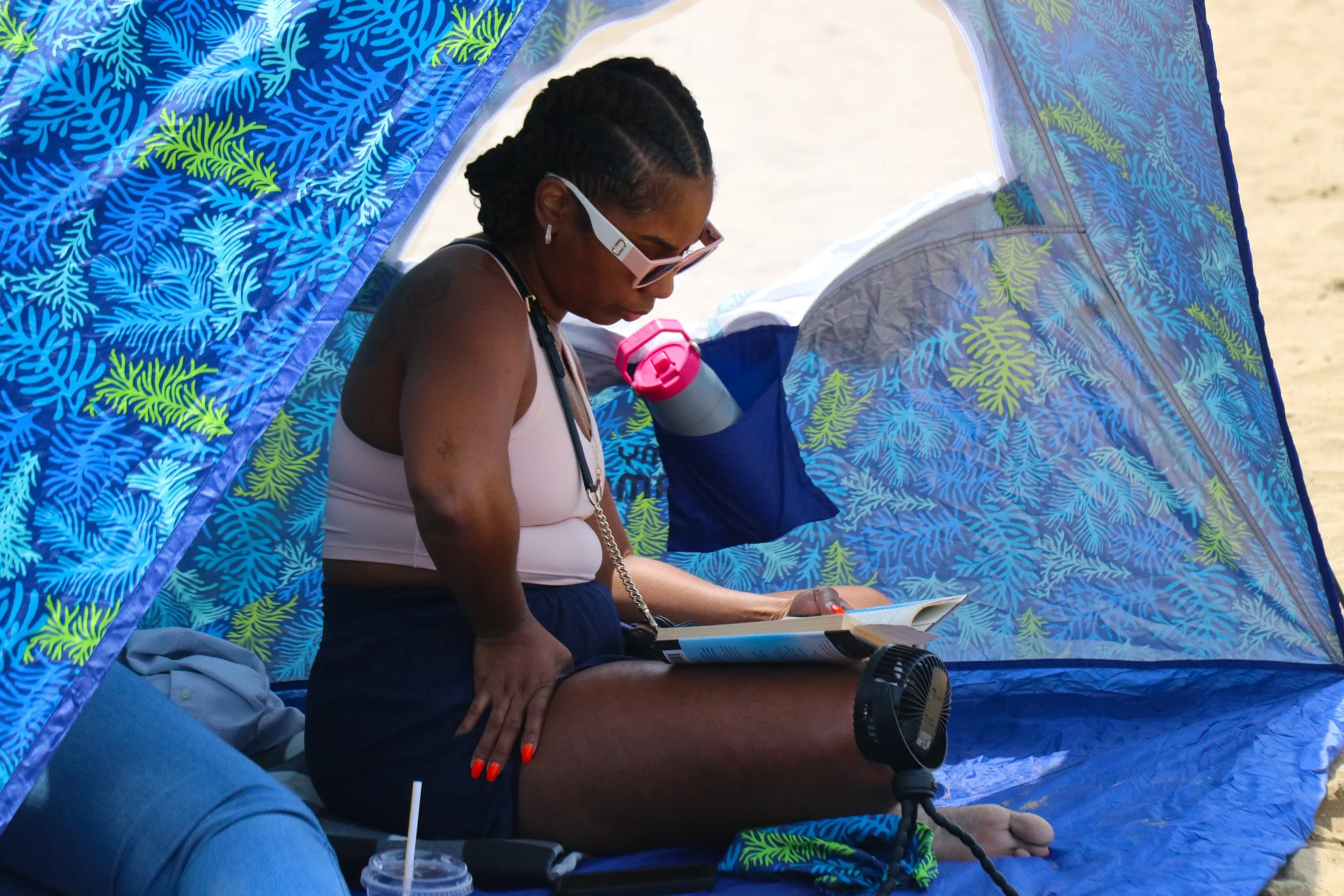 A woman sitting inside a blue beach tent reading a book, with a small fan in front of her, near the sand.