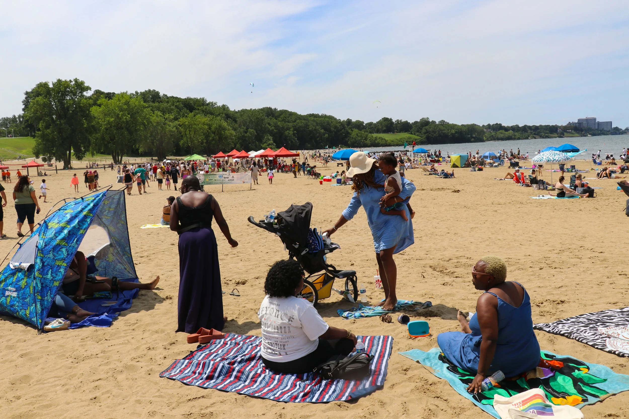 People relaxing and socializing on a sunny beach near a body of water, with tents, umbrellas, and trees in the background.