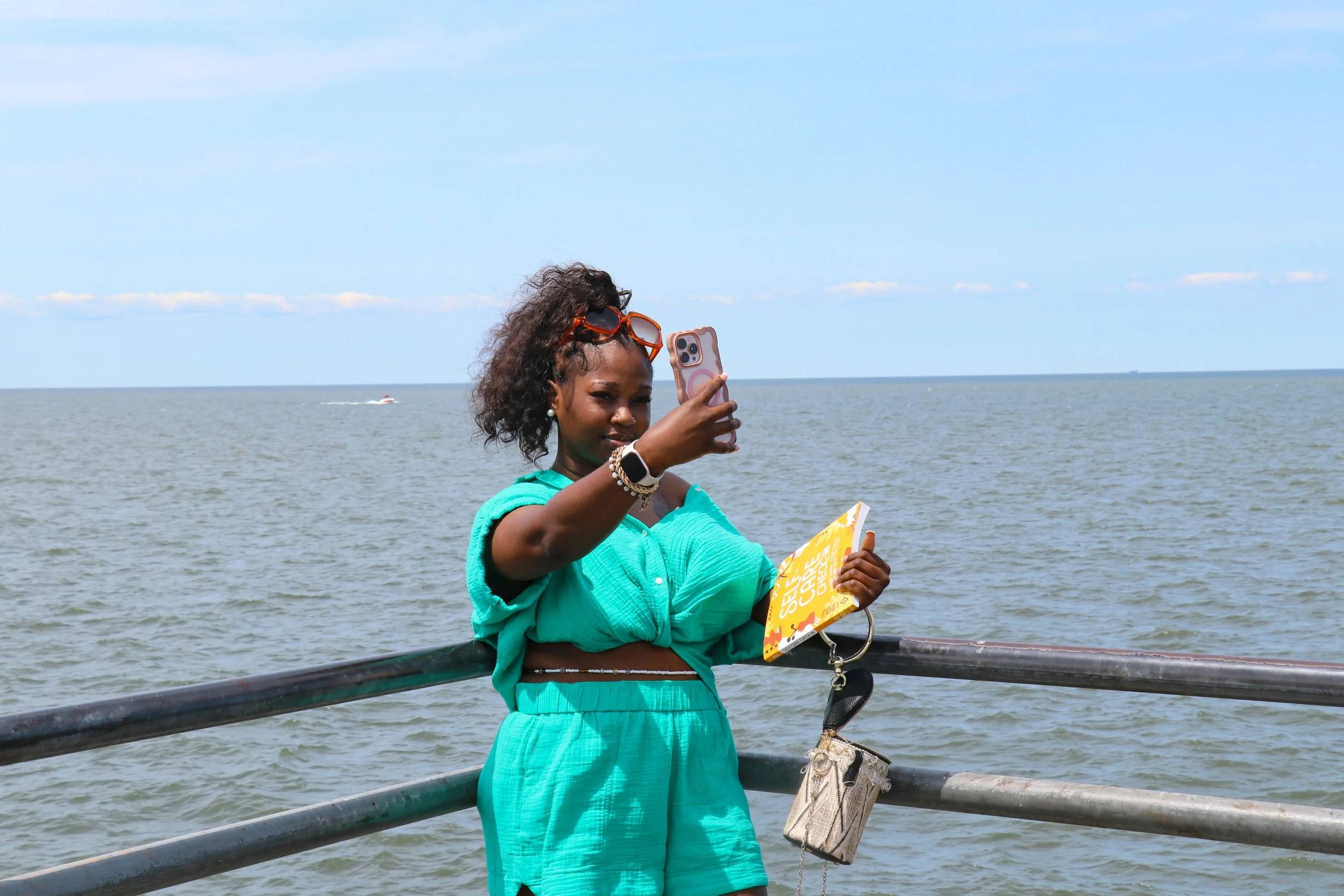 A woman in a teal outfit taking a selfie by the water at a seaside location.