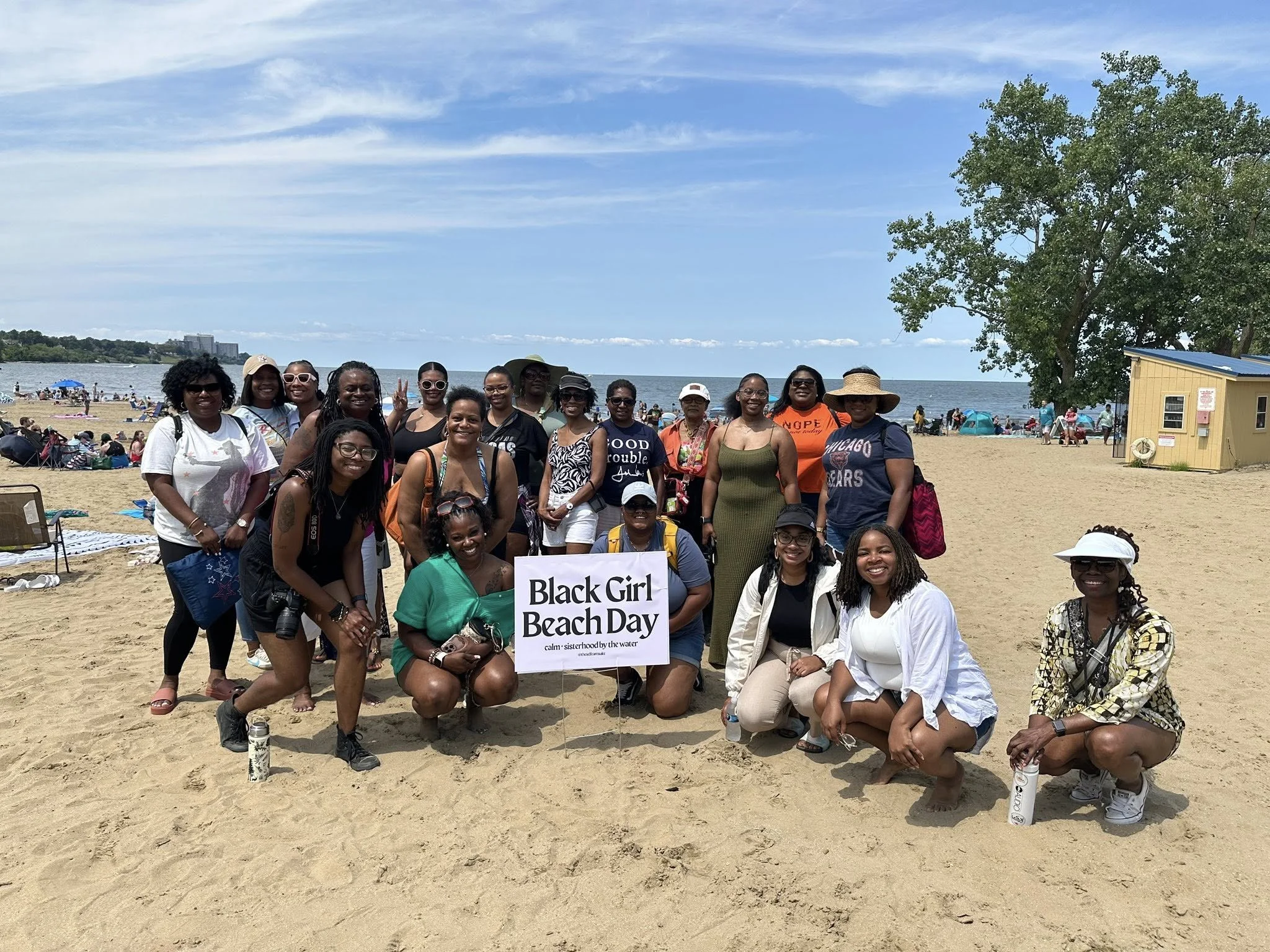 Group of women and girls on a sandy beach holding a sign that reads "Black Girl Beach Day" with the water and a few buildings in the background.