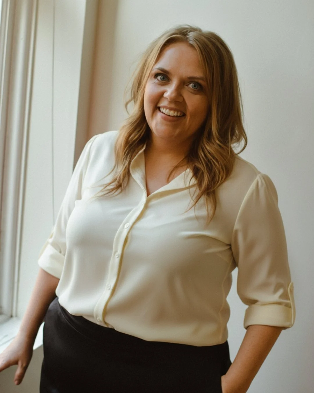 Heather Roberts, skincare specialists at Mode Beauty NYC. A woman with shoulder-length wavy light brown hair and blue eyes smiling, wearing a cream button-up shirt with rolled-up sleeves, standing indoors near a window with light-colored curtains.