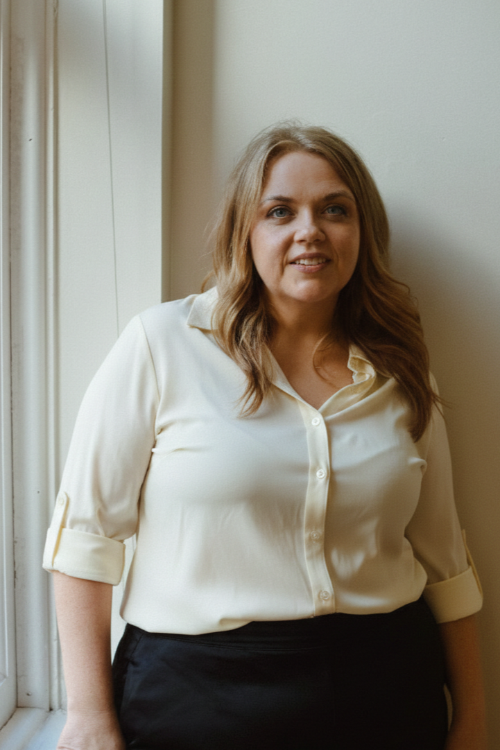Heather Roberts, licensed esthetician and owner at Mode Beauty NYC, with shoulder-length light brown hair, wearing a cream-colored blouse with rolled-up sleeves and black pants, standing by a window with a neutral wall background.
