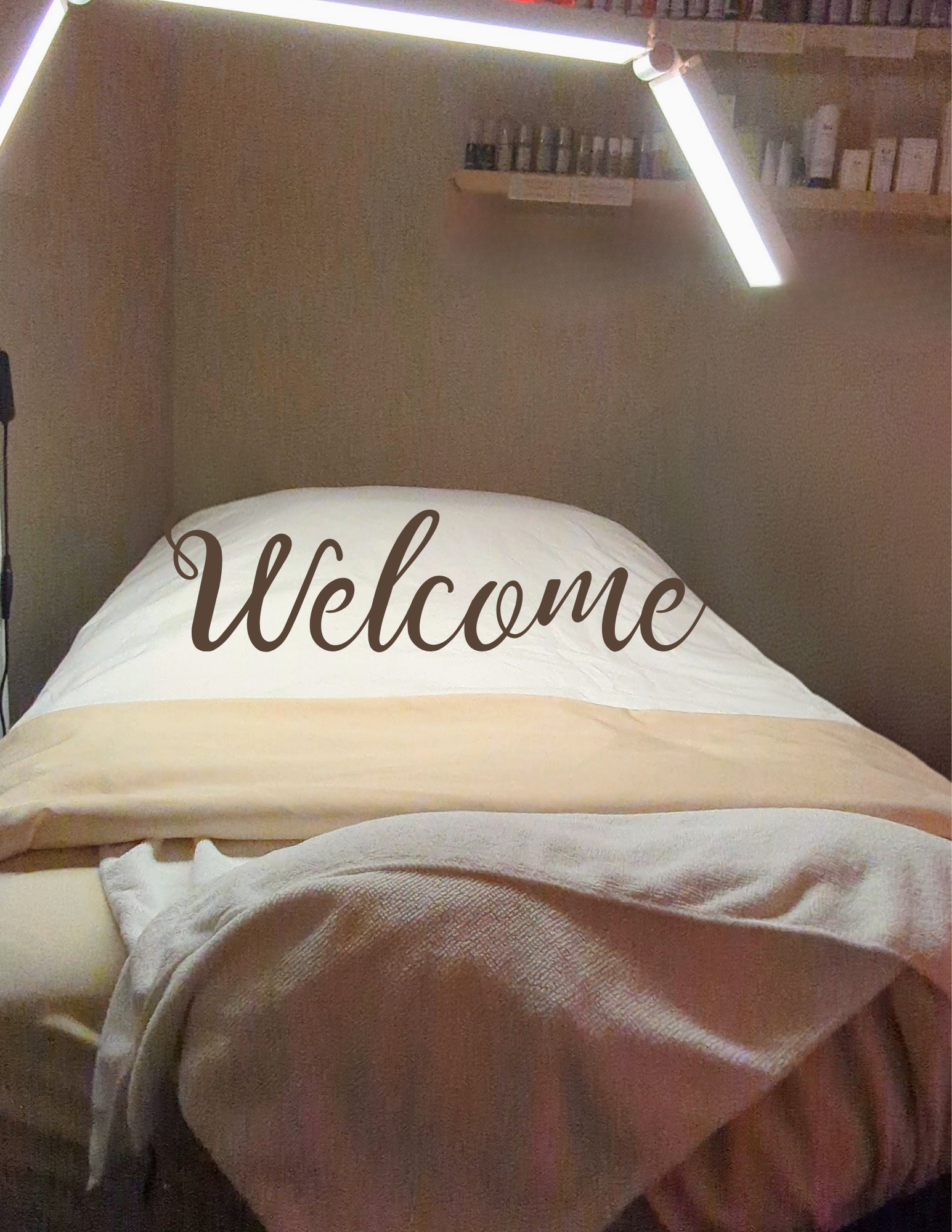 A neatly made bed with a pillow that has the word 'Welcome' written on it, in a cozy bedroom with a book shelf and modern lighting.