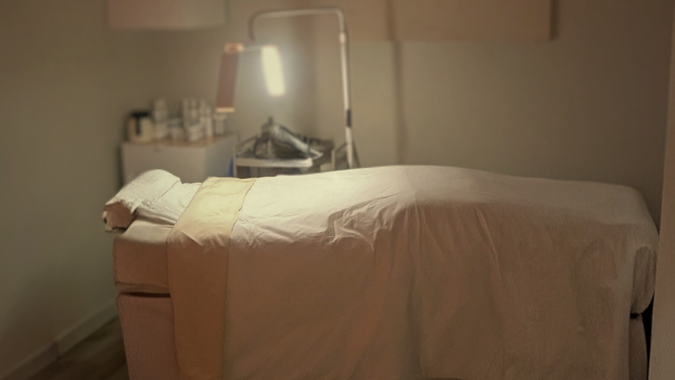 An image of the treatment room at Skincare By Heather in Flatiron NYC. It shows a treatment table set up for a facial with a pillow and blanket. Products are in the background and a facial lamp is glowing