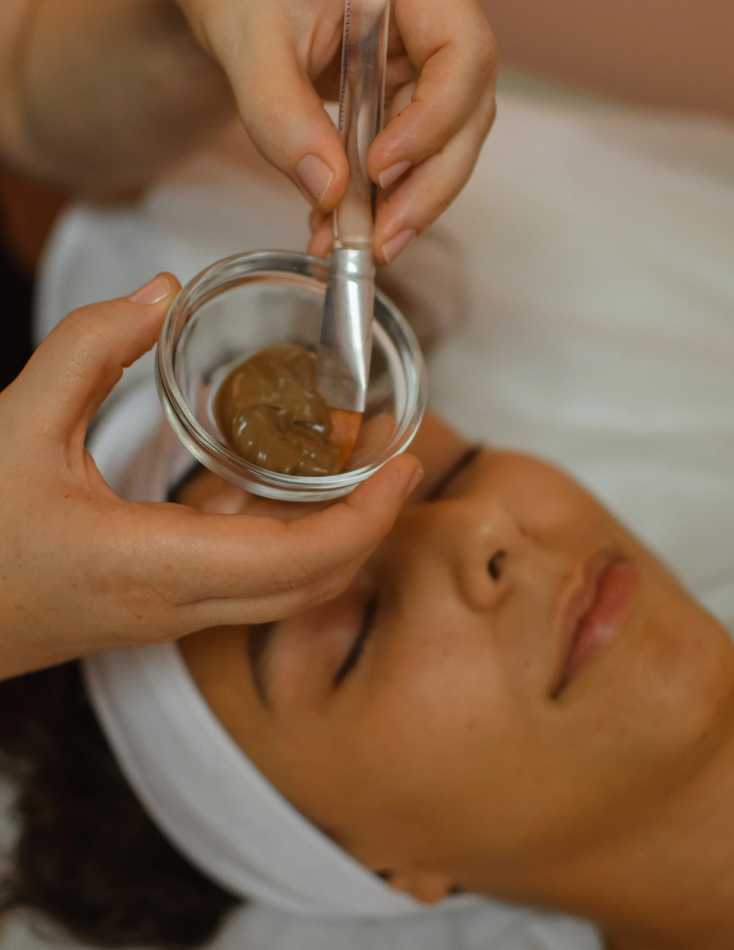 A person receiving a facial treatment with a small glass container filled with a brown mask or cream, applied on their face, while lying down with eyes closed and head wrapped with a white towel.
