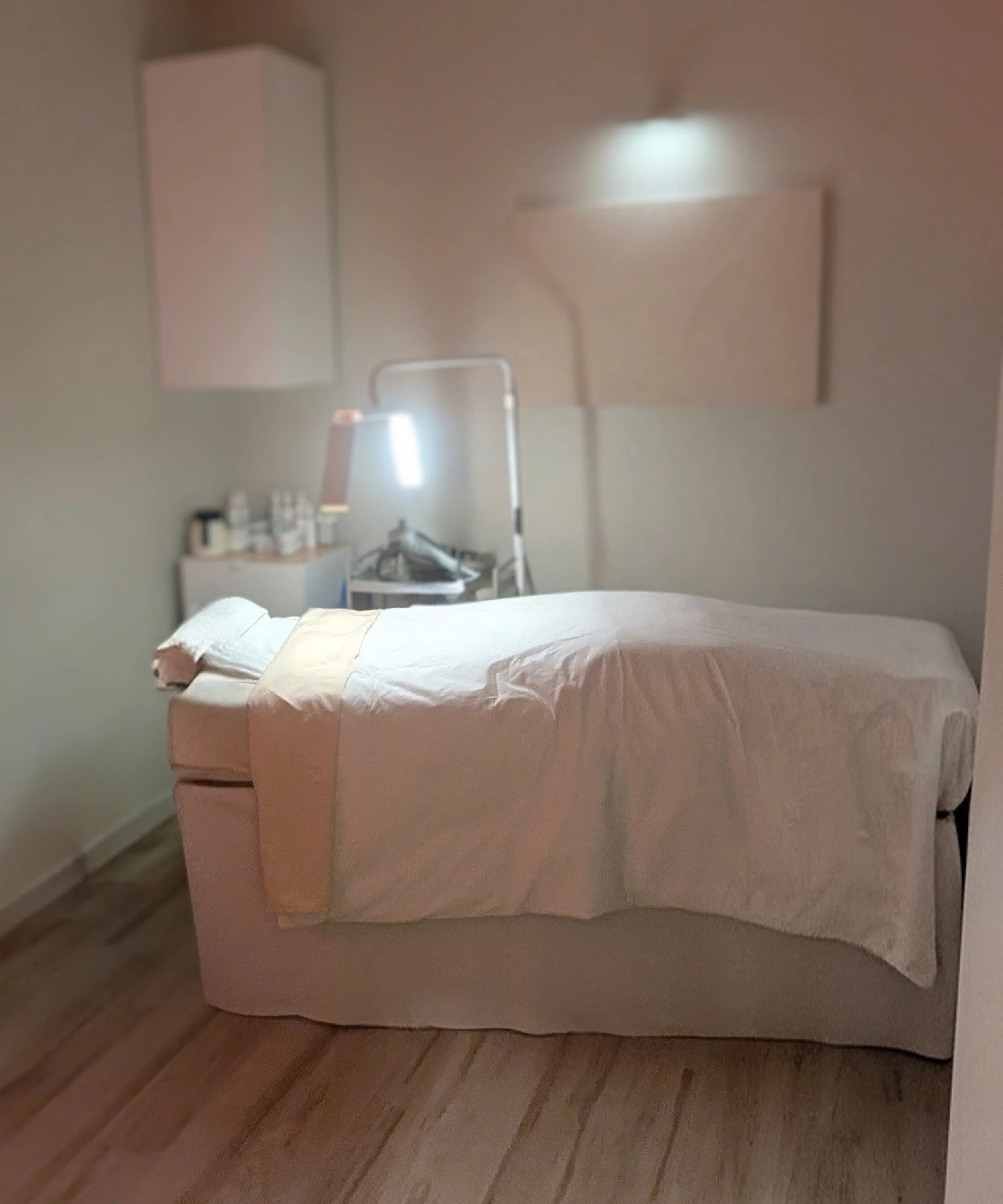A massage or treatment room with a bed covered in white linens, a light on a stand, and shelves with bottles and supplies in the background.