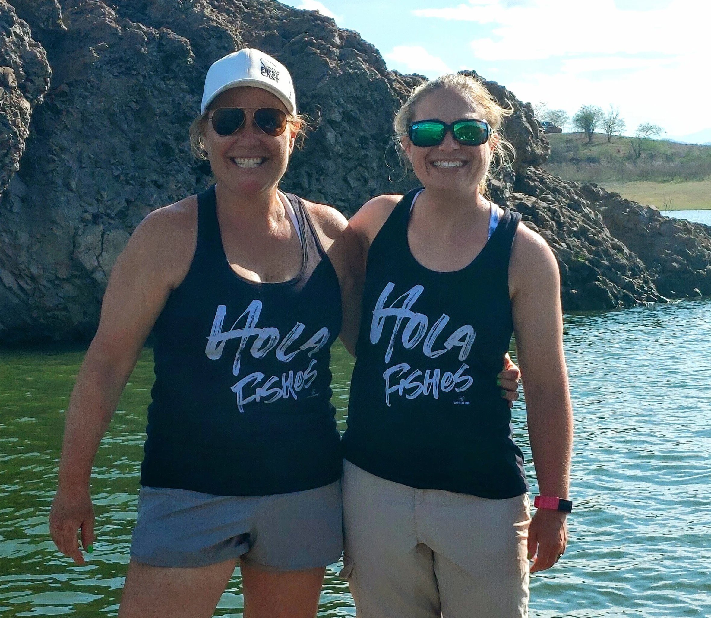 Two women standing together on a water body, wearing black tank tops with 'HOLA FISHES' written on them, smiling, with rocky hills and a partly cloudy sky in the background.