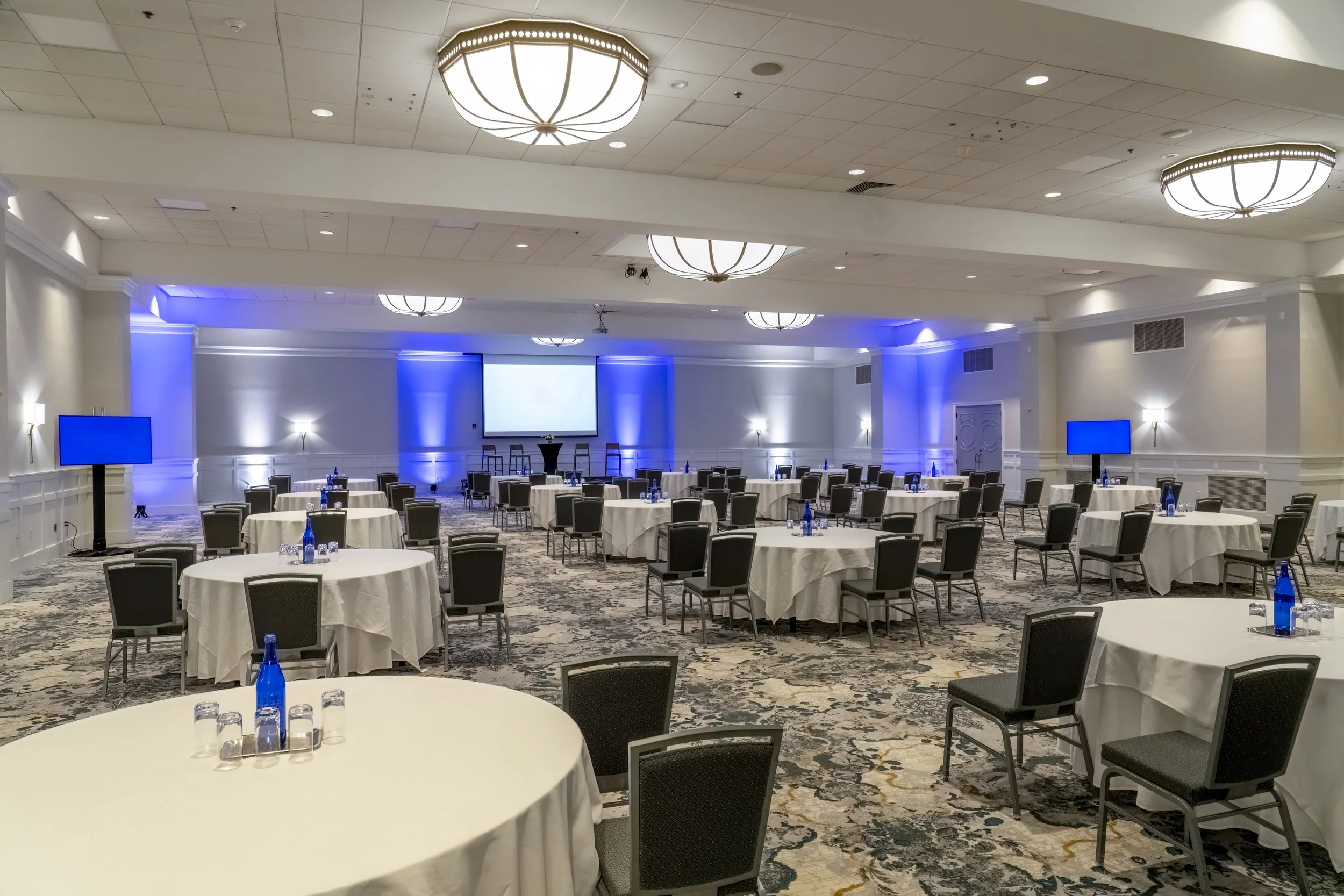 Conference room with round tables covered with white tablecloths, each set with water bottles and glasses, facing a stage with a large screen and blue lighting.