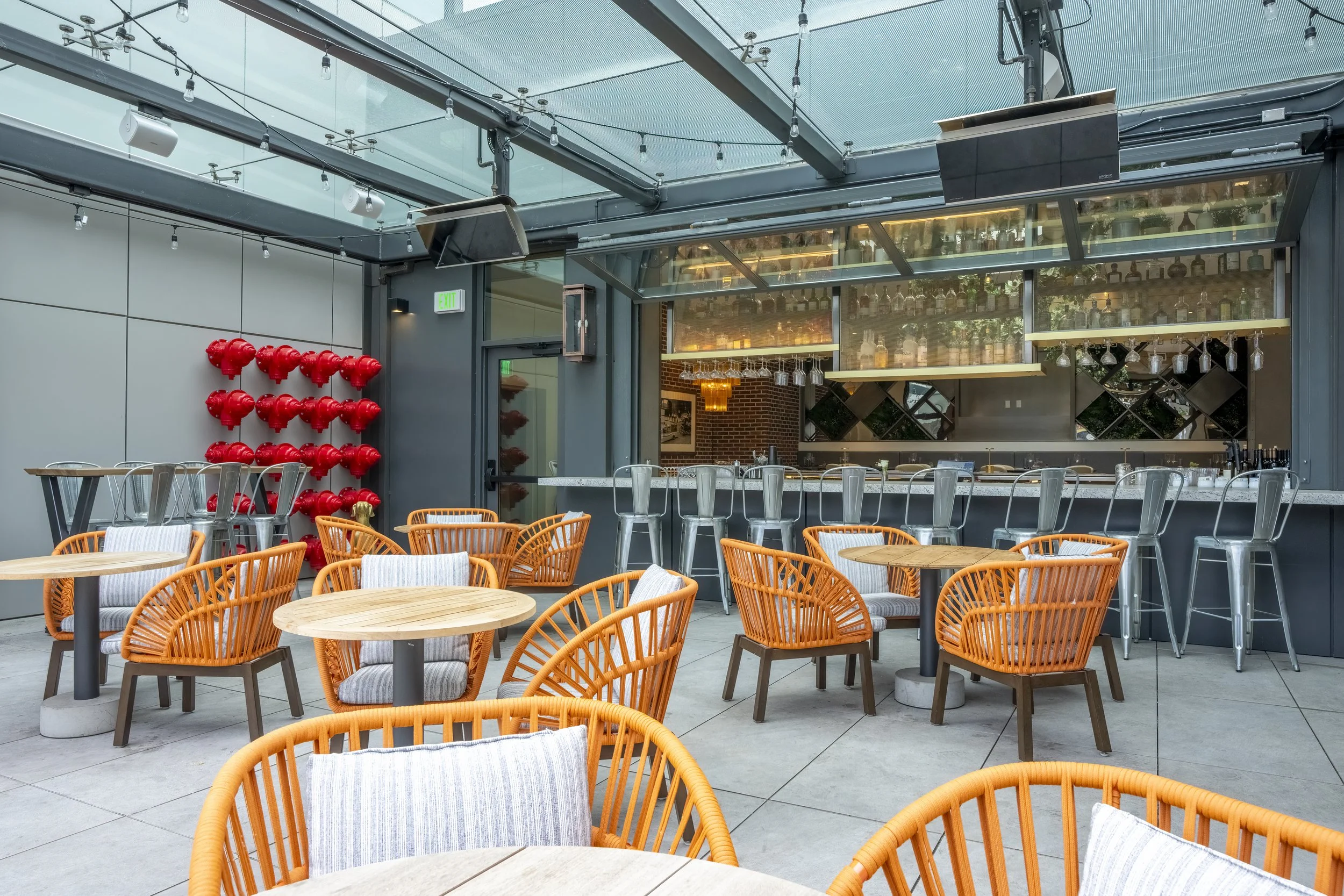 An outdoor patio area of a bar or restaurant with round wooden tables and orange chairs with striped cushions. There is a bar with a row of metal bar stools and hanging glasses. A wall on the left has red fire hydrant decorations. The space is partially covered with string lights overhead.