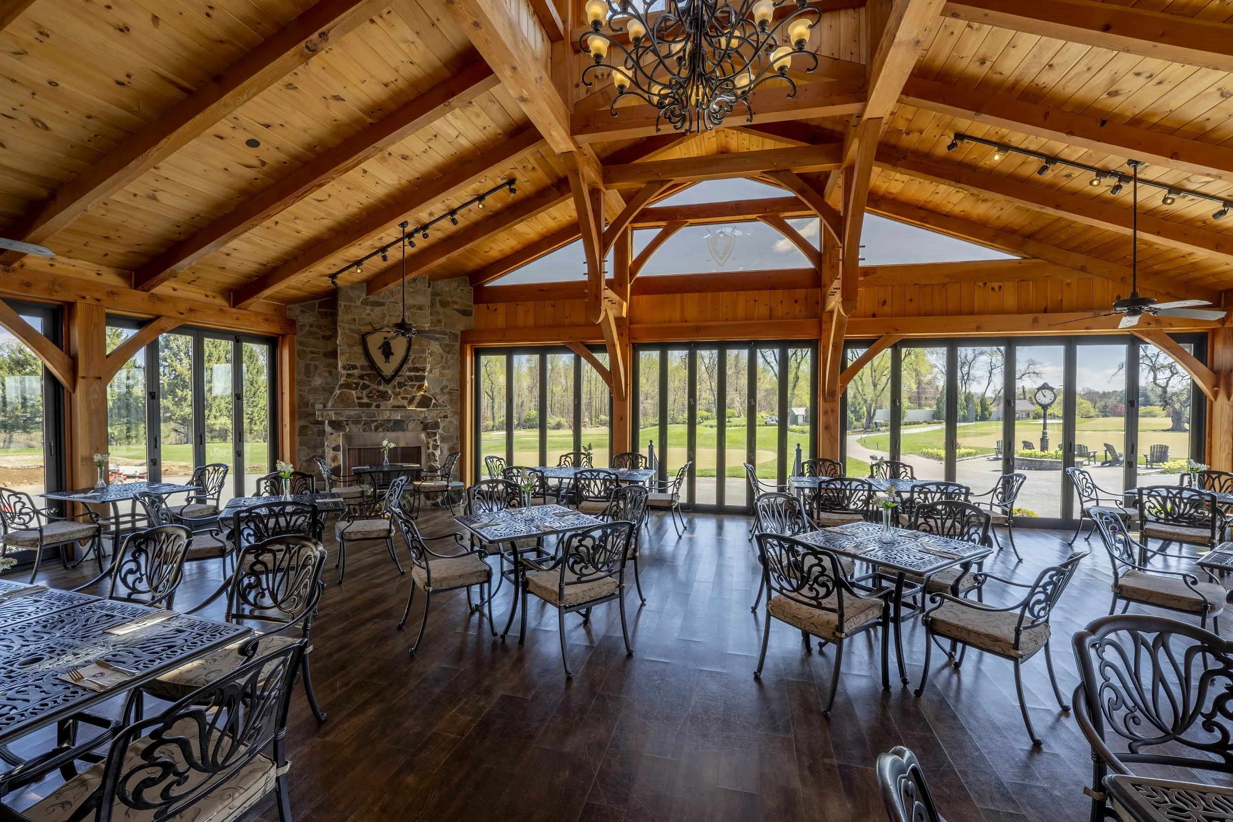 Inside of a spacious event hall with a high wooden vaulted ceiling, large windows showing a green outdoor landscape, tables and chairs set up for dining, a stone fireplace, and chandeliers hanging from the ceiling.