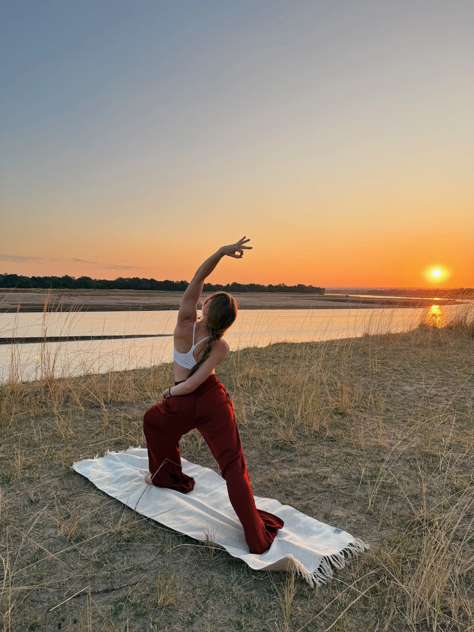 A woman practicing yoga on a towel outdoors at sunset near a body of water, kneeling with one arm extended upward and the other hand placed on her waist, surrounded by dry grasses.