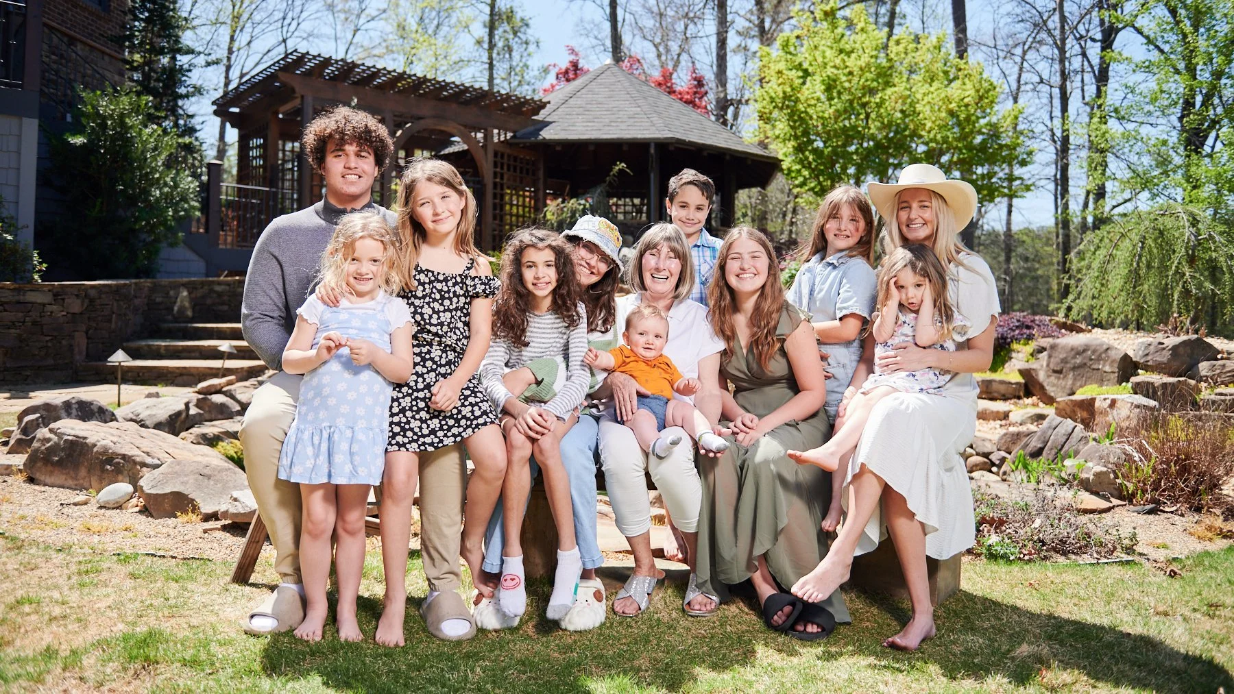 Large multi-generational family group with children, teenagers, and adults gathered around the matriarch grandmother outdoors for a family reunion portrait.