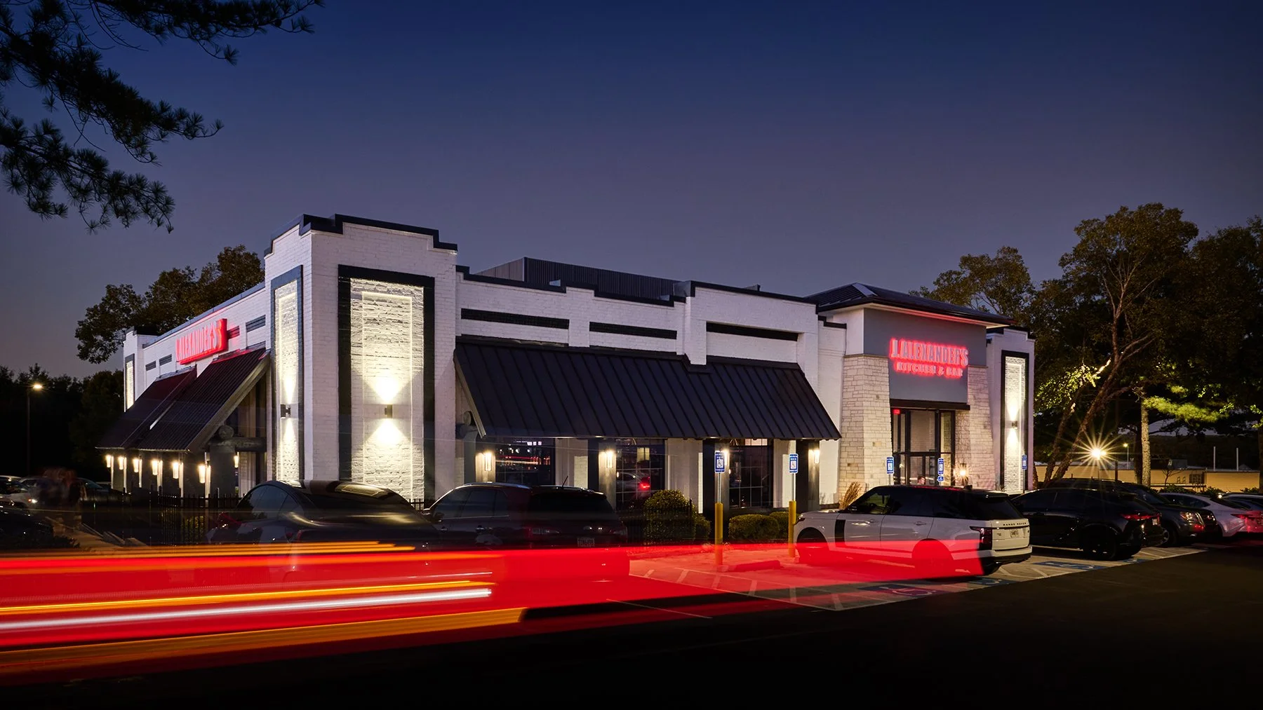 Twilight architectural photography of J. Alexander's restaurant with illuminated exterior, vehicle light trails, and evening activity in Peachtree City, Georgia.