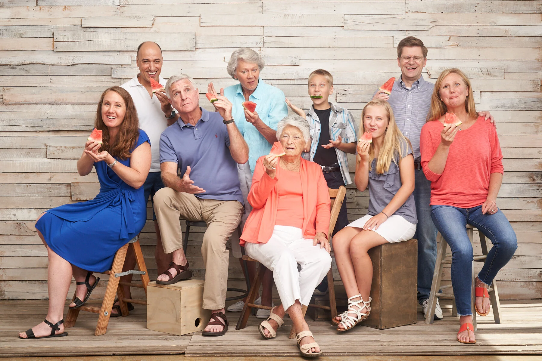 Multi-generational family laughing and eating watermelon together during a playful indoor family portrait session against a light wood backdrop.