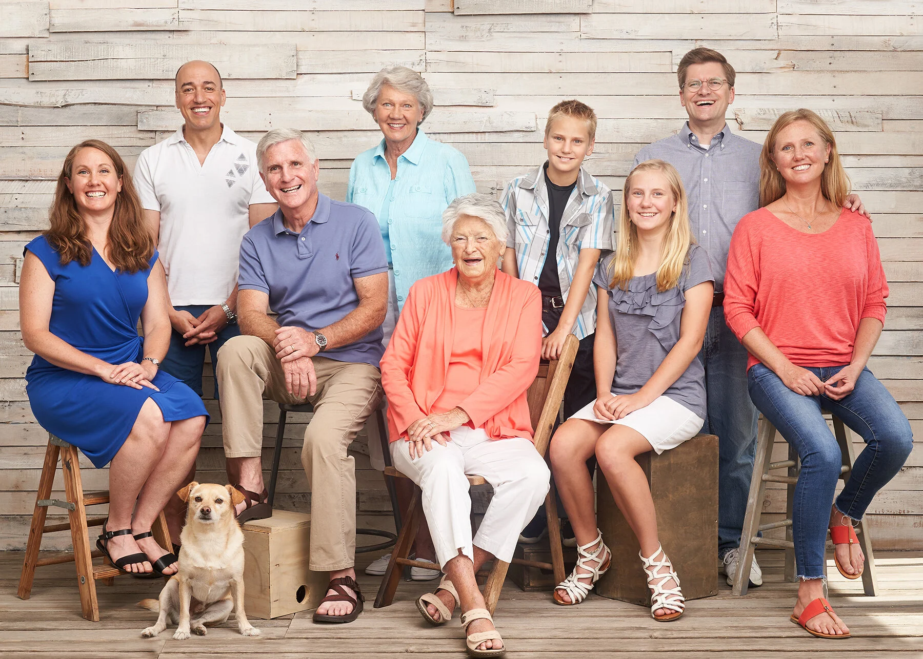 Multi-generational family of nine adults and children posed together with their dog during an indoor family portrait session against a light wood backdrop.