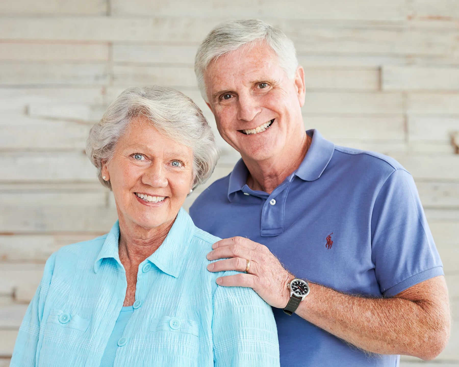 Older married couple smiling together during a relaxed indoor family portrait session against a light wood backdrop.