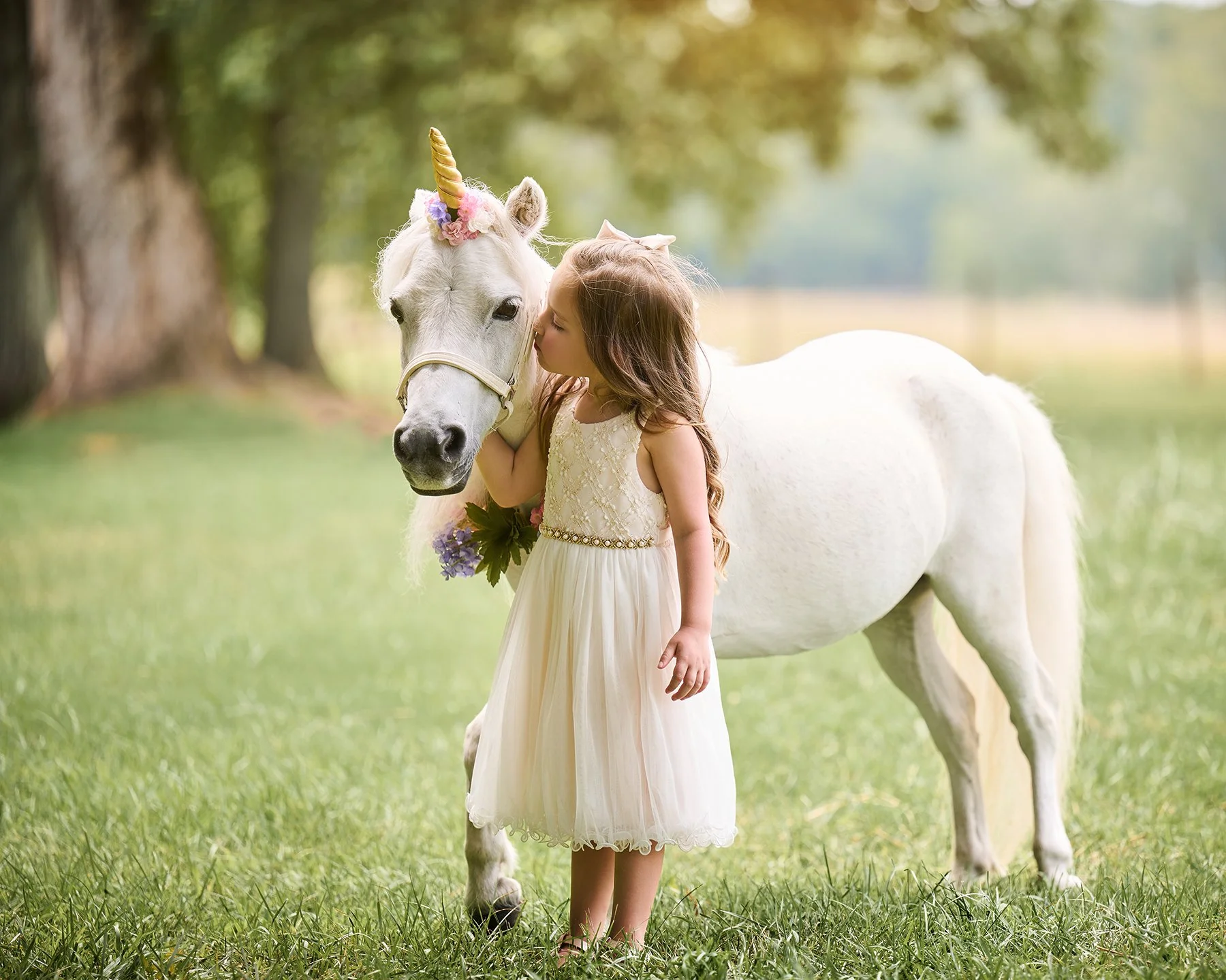 Five-year-old girl in a white dress gently kissing a white pony styled as a unicorn with pastel flowers and a gold horn during an outdoor portrait session in South Atlanta.