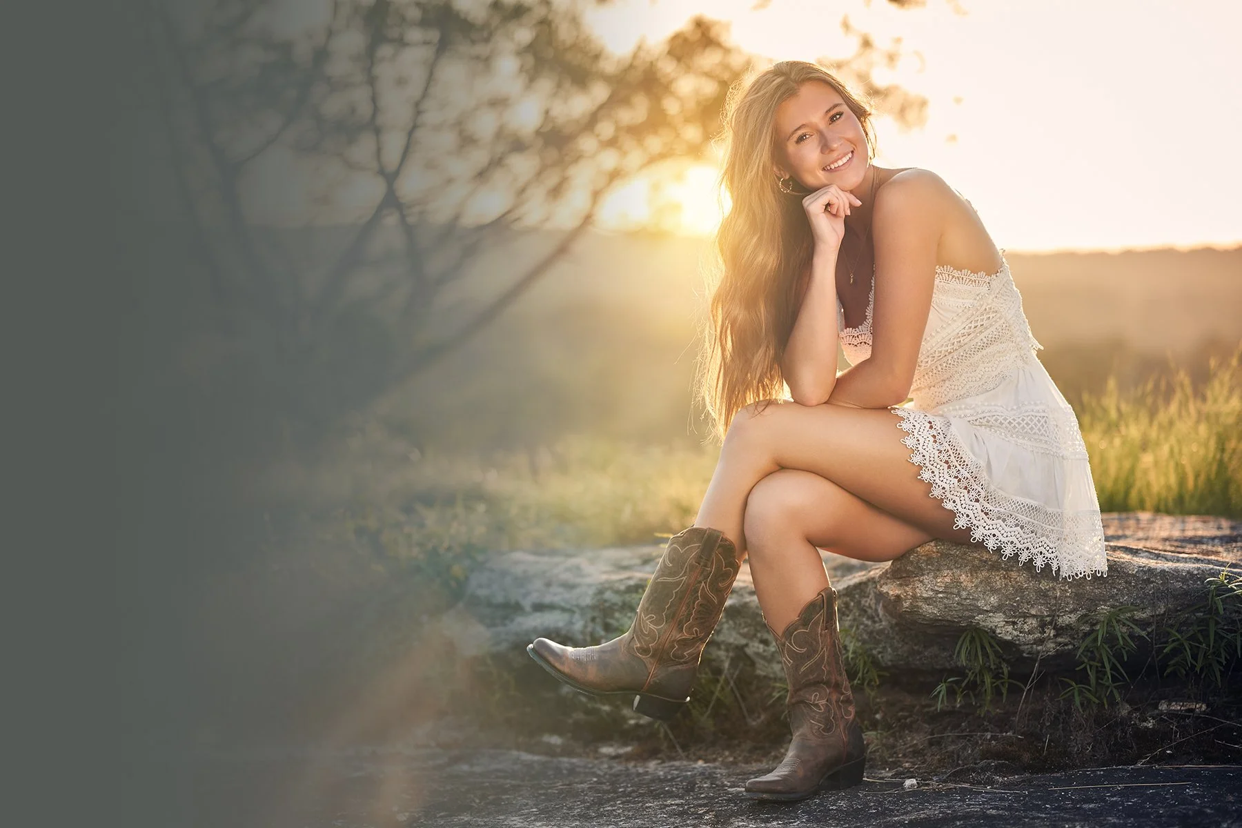 Senior girl in a white lace dress and cowboy boots seated on a rock in warm golden-hour light during an outdoor senior portrait session in Peachtree City, Georgia.
