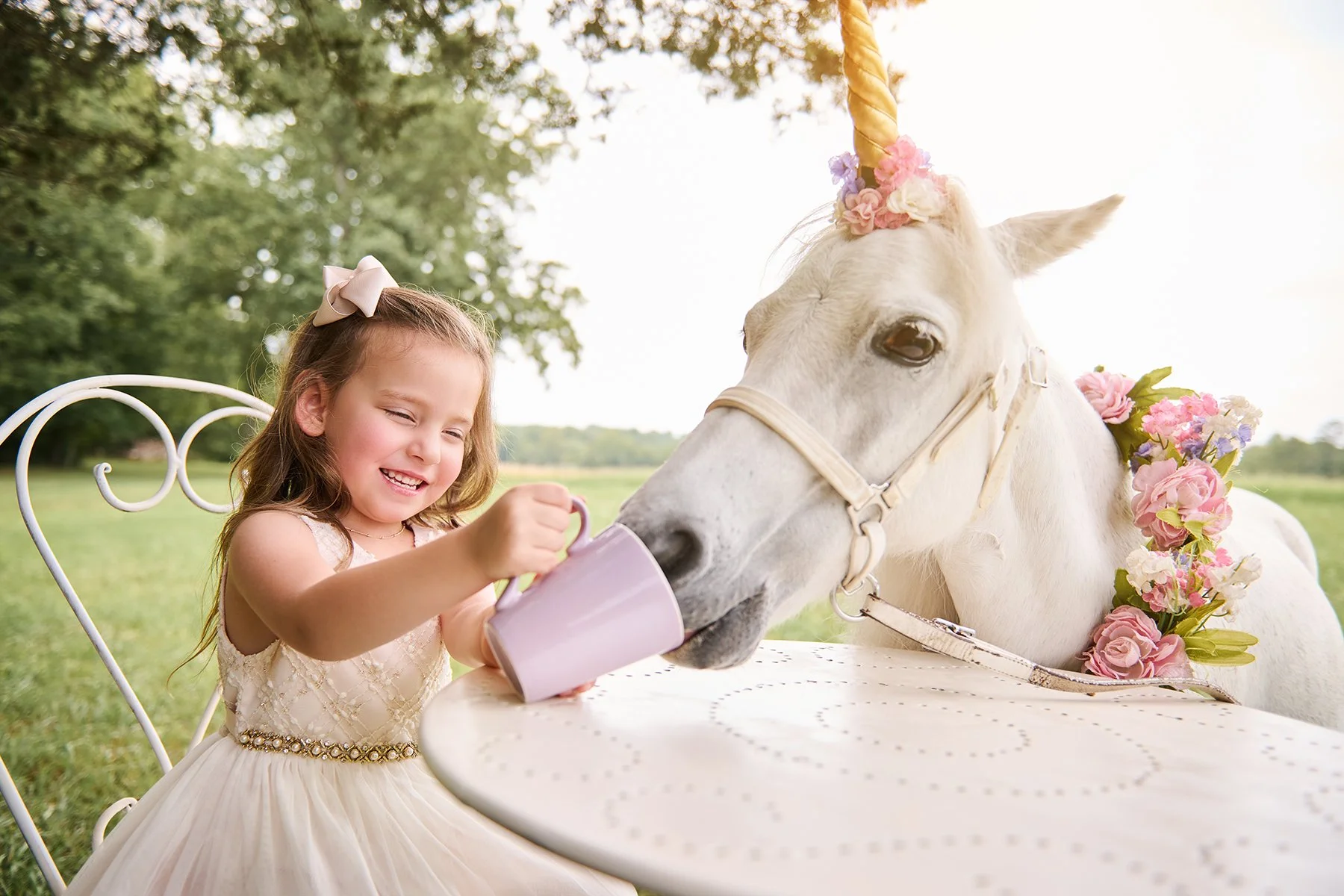 Five-year-old girl in a white dress pretending to serve tea to a white unicorn with pastel flowers and a gold horn during an outdoor birthday portrait session.