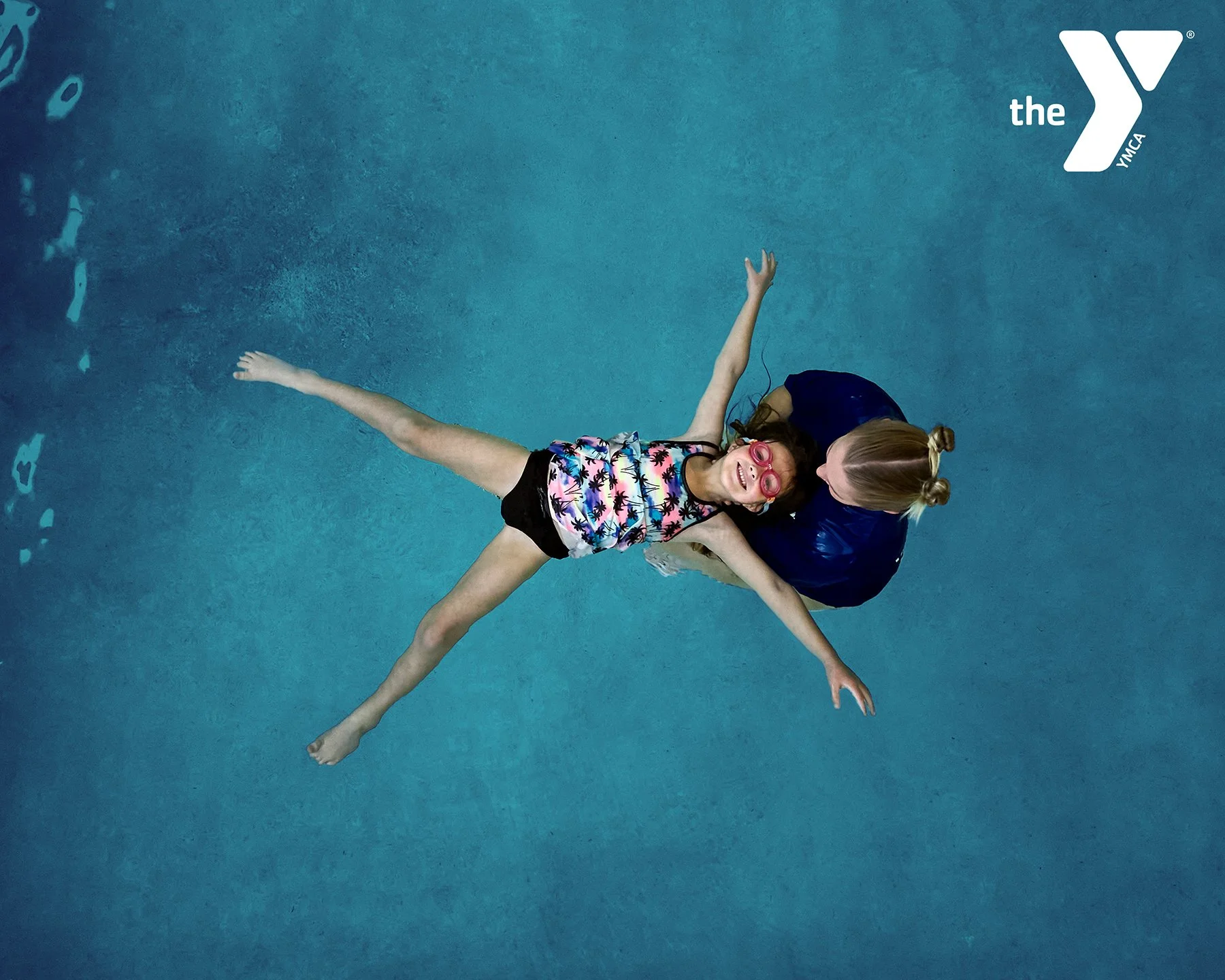 Overhead drone photography of a YMCA swim instructor supporting a young swimmer in a blue pool, captured for a commercial marketing campaign.