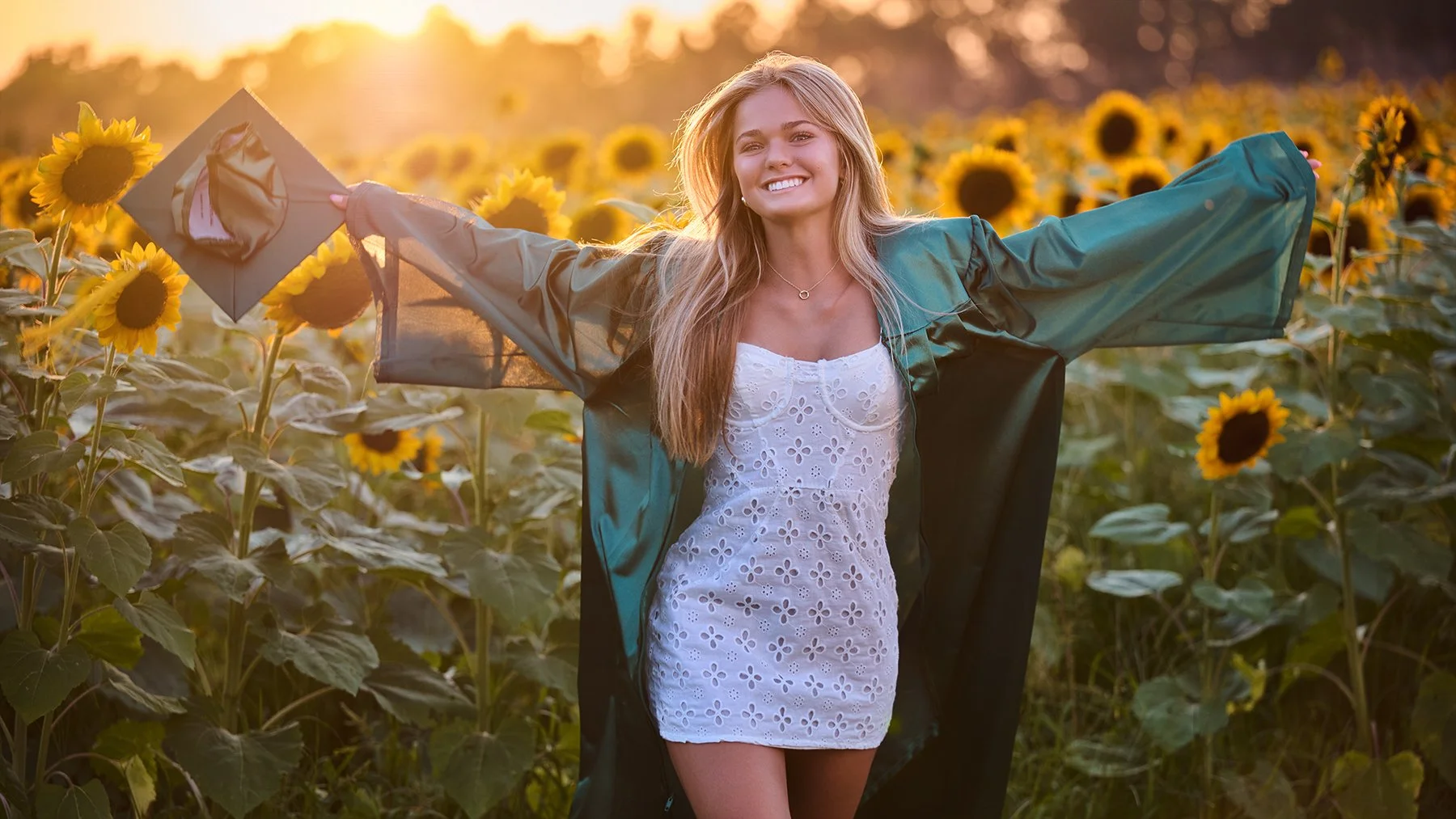 Outdoor senior portrait of a smiling blonde graduate wearing a white dress and green graduation gown, standing with her arms extended in a sunflower field at sunset.