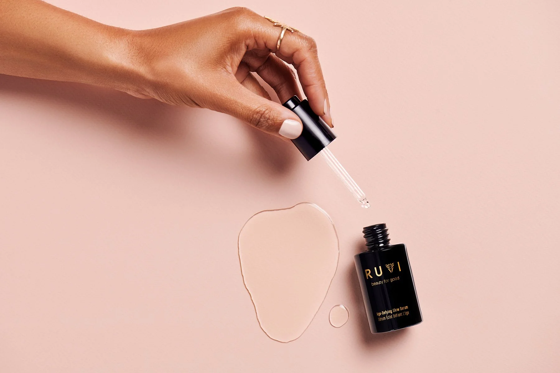 Studio product photography of RUVI Age-Defying Glow Serum with glass dropper and liquid texture against a soft pink background for a beauty marketing campaign.