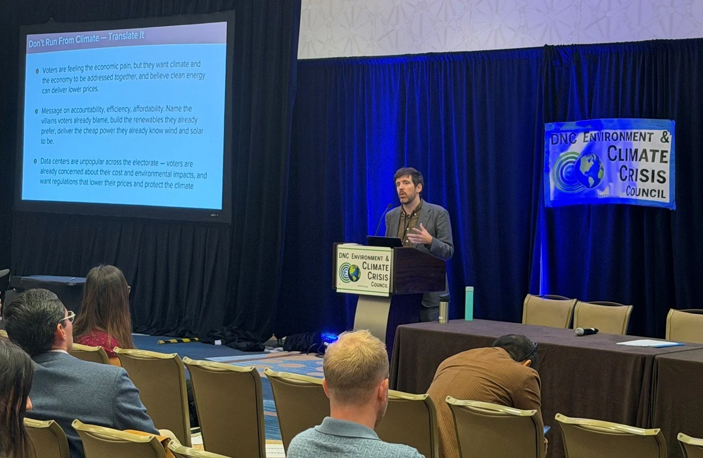 Ryan O'Donnell speaks and gestures at a podium in front of a projection screen reading "Don't Run From Climate — Translate It," with a "DNC Environment & Climate Crisis Council" banner beside him.