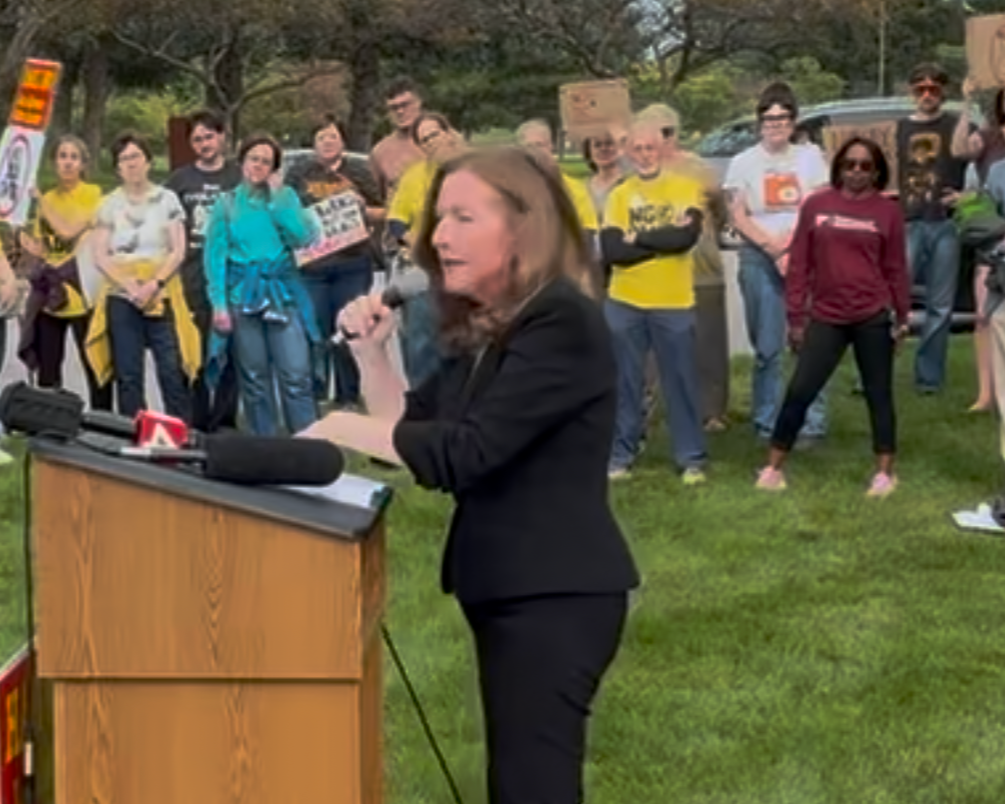 DNC Climate Council Chair Michelle Deatrick, wearing a black suit, holds a microphone and stands at a wood podium. A crowd of protestos in colorful yellow, blue and white clothes stand behind her.