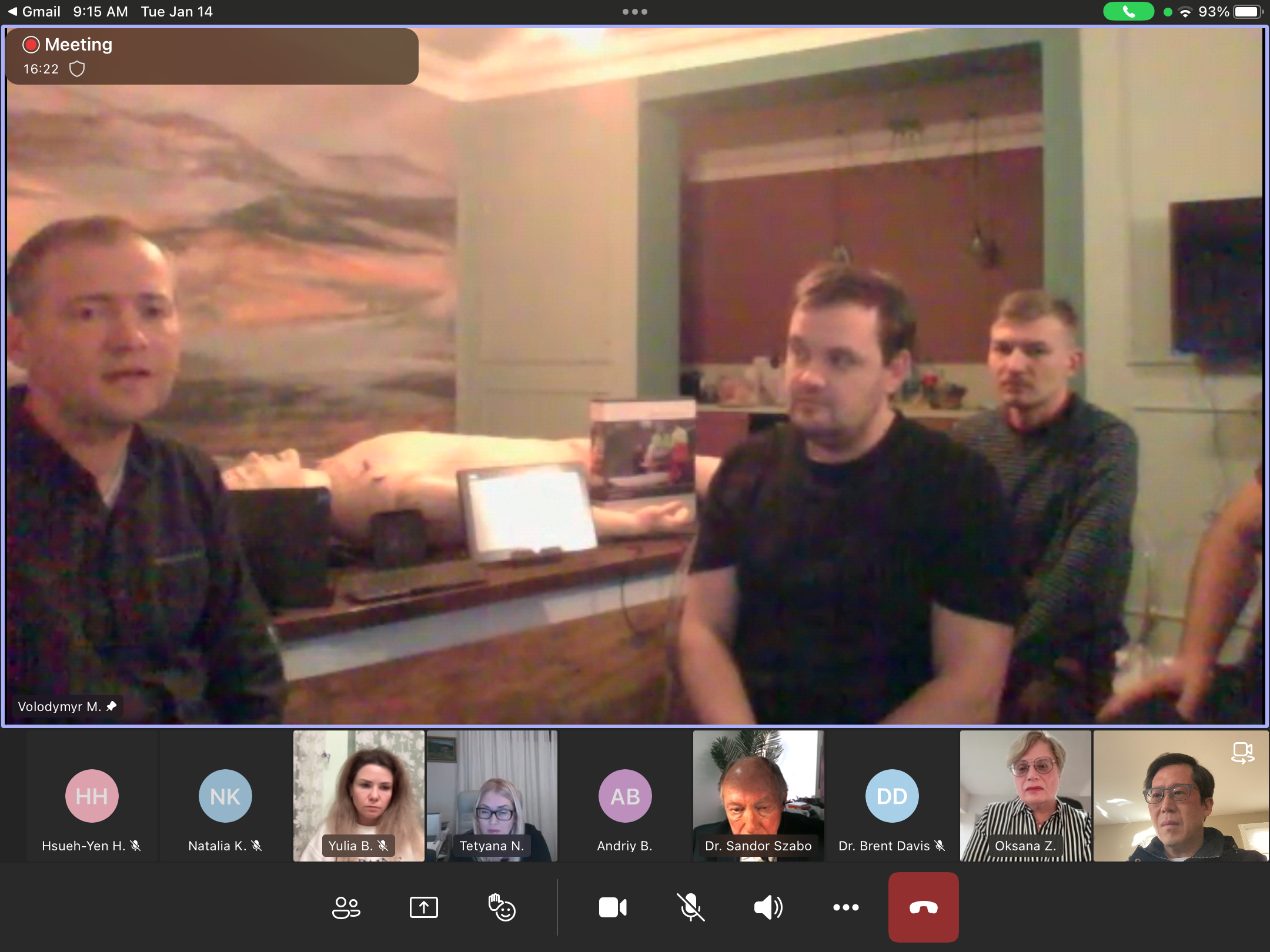 A screenshot of a virtual meeting with multiple participants. The main window shows three men sitting in a room with a wooden bar, some bottles, and a TV mounted on the wall. The virtual participants are displayed at the bottom, with their names and 