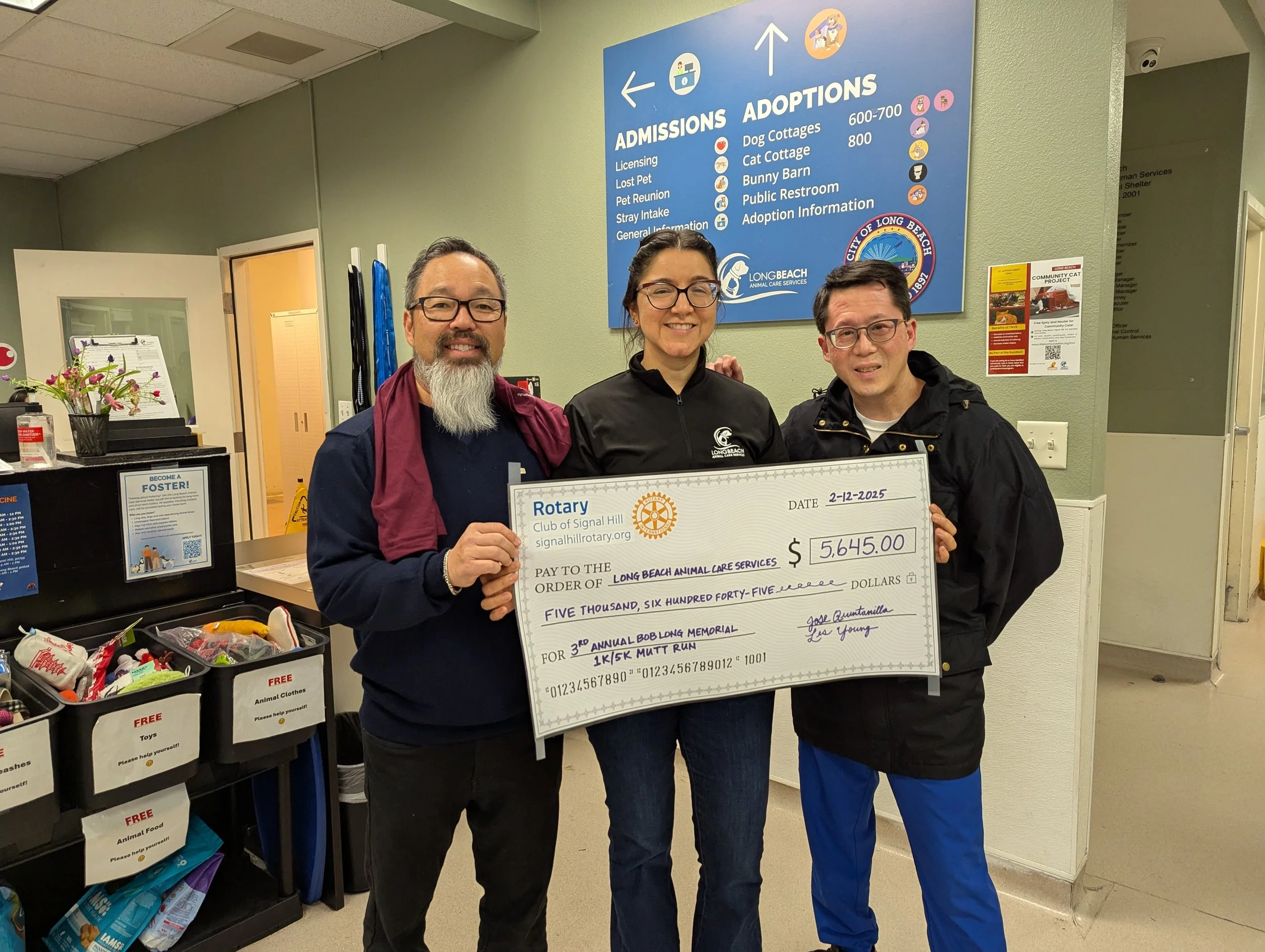 Three people standing indoors, holding a large check made out to Long Beach Animal Care Services for $5,645, at a donation event. One person is a man with glasses and a beard, another is a woman with glasses and dark hair, and the third is a man with glasses and dark hair. They are smiling and standing in front of a blue sign with directions and information about animal adoptions and admissions.