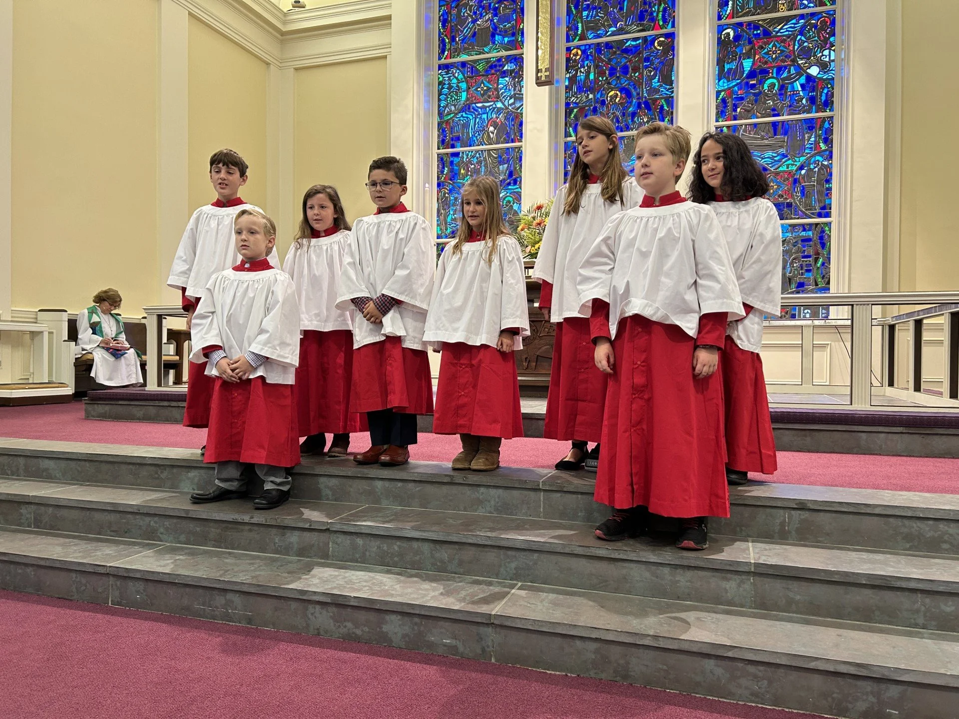 Children in choir robes performing in a church with stained glass windows in the background.