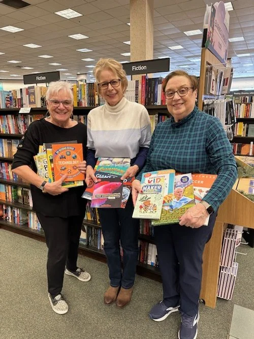Three women standing in a bookstore aisle holding kids' craft and technology books, with shelves labeled 'Fiction' behind them.