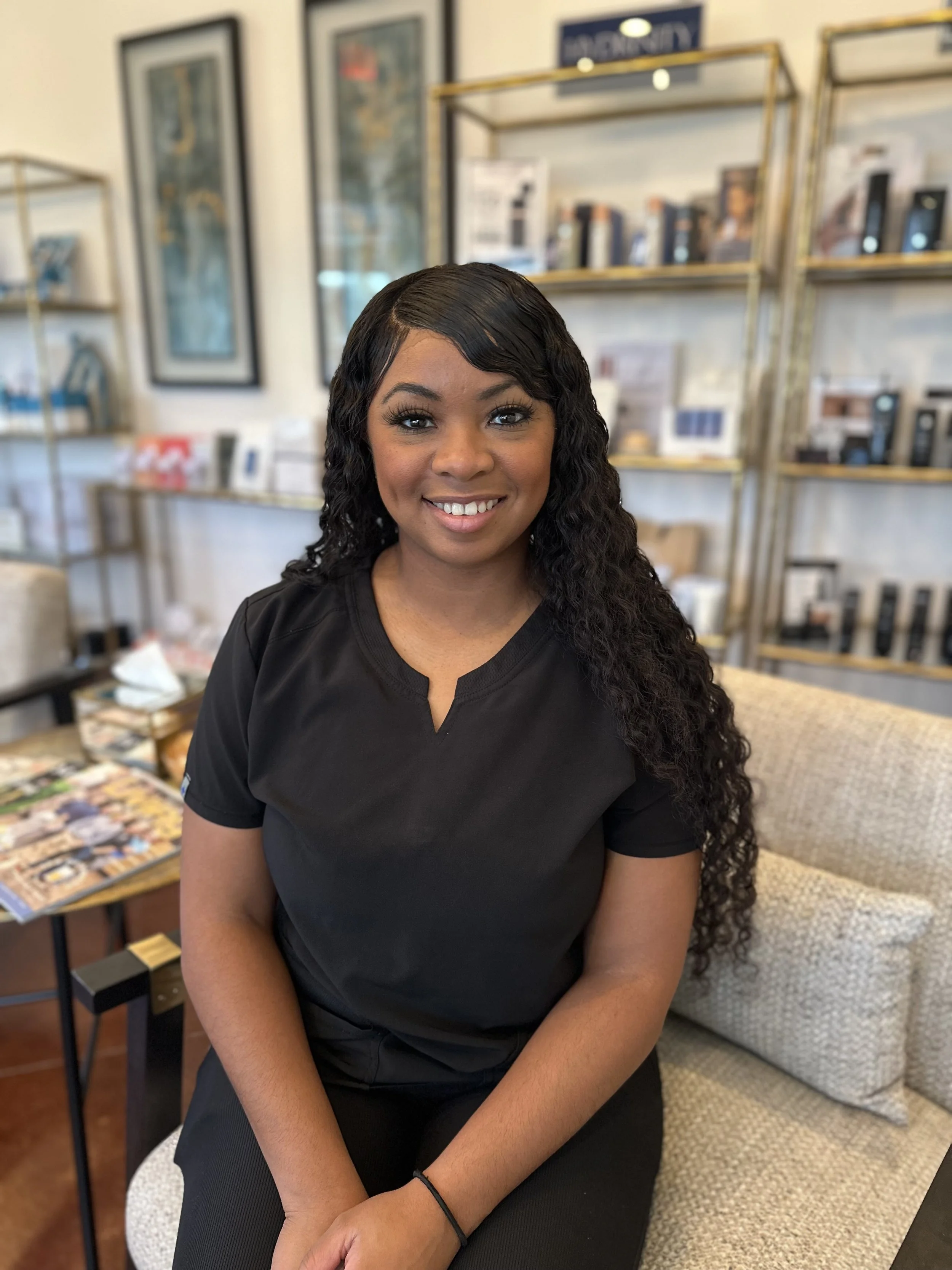 A smiling woman with long curly black hair sitting in a casual setting with framed pictures and shelves in the background, wearing a black top.