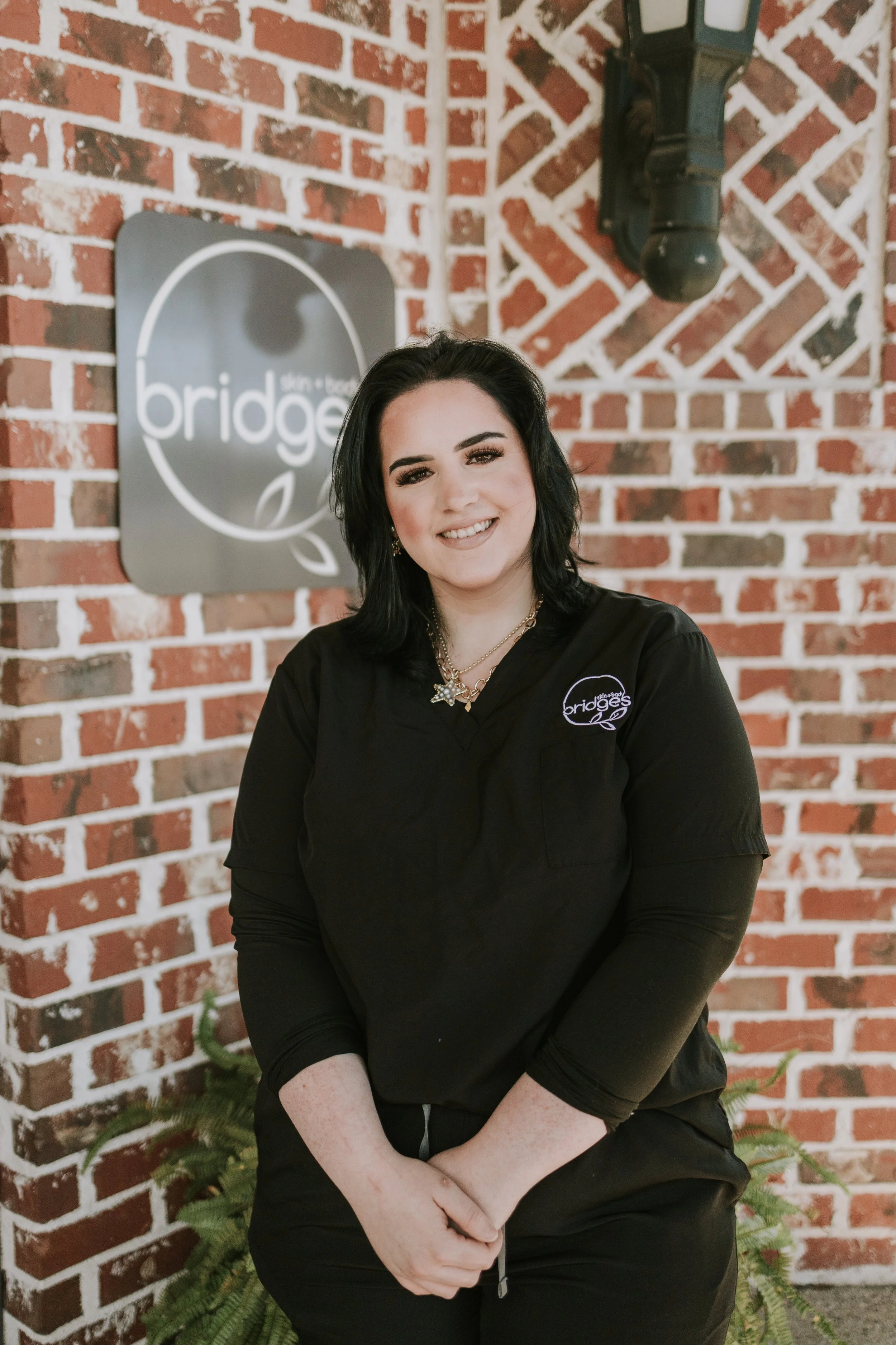 A woman with black hair, smiling, wearing a black shirt with a logo that says 'bridges,' sitting in front of a brick wall with a sign of the same name and a black pipe above her head. Potted ferns are at her sides.