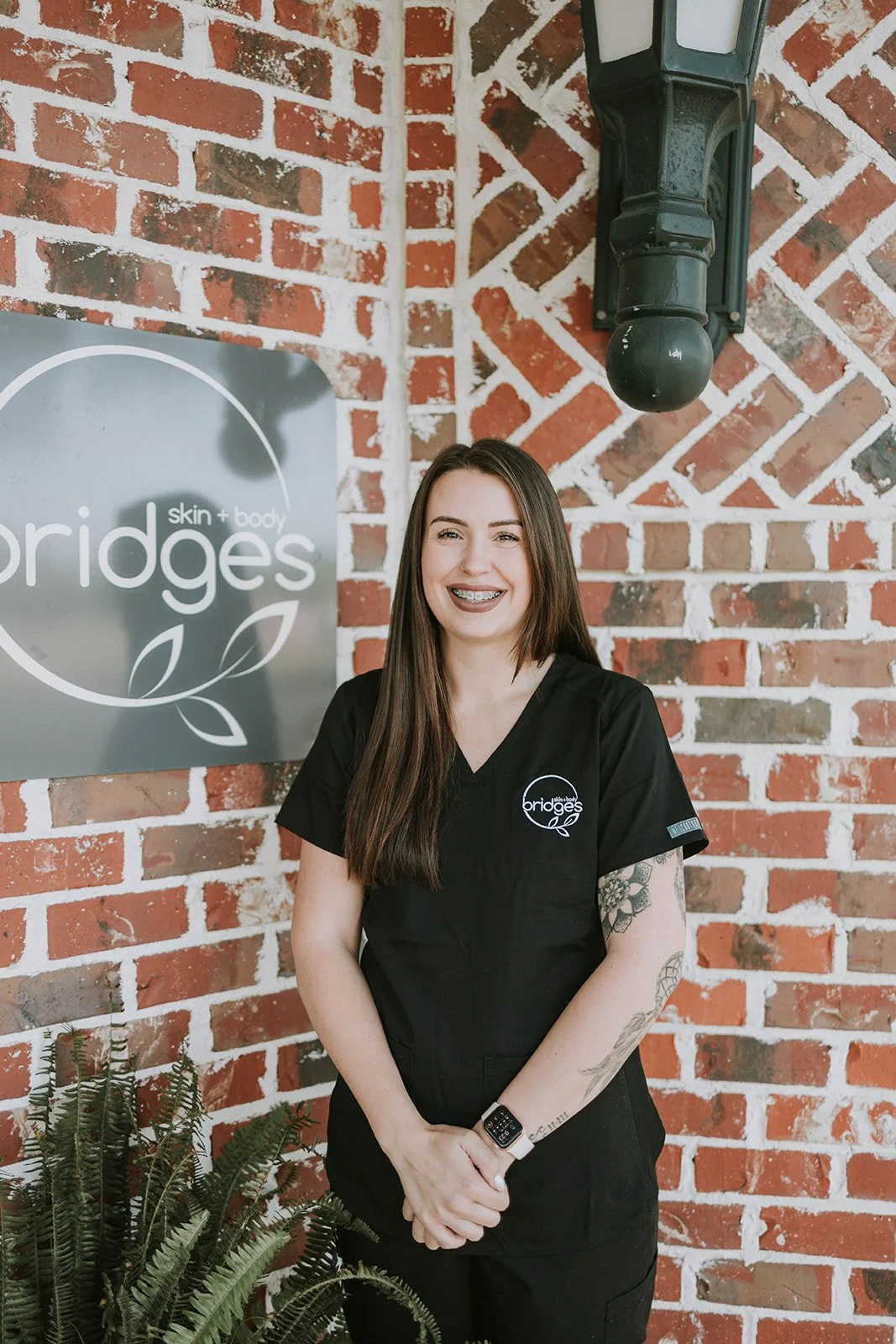 A woman with long brown hair, dressed in black scrubs, smiling, standing in a skincare store.