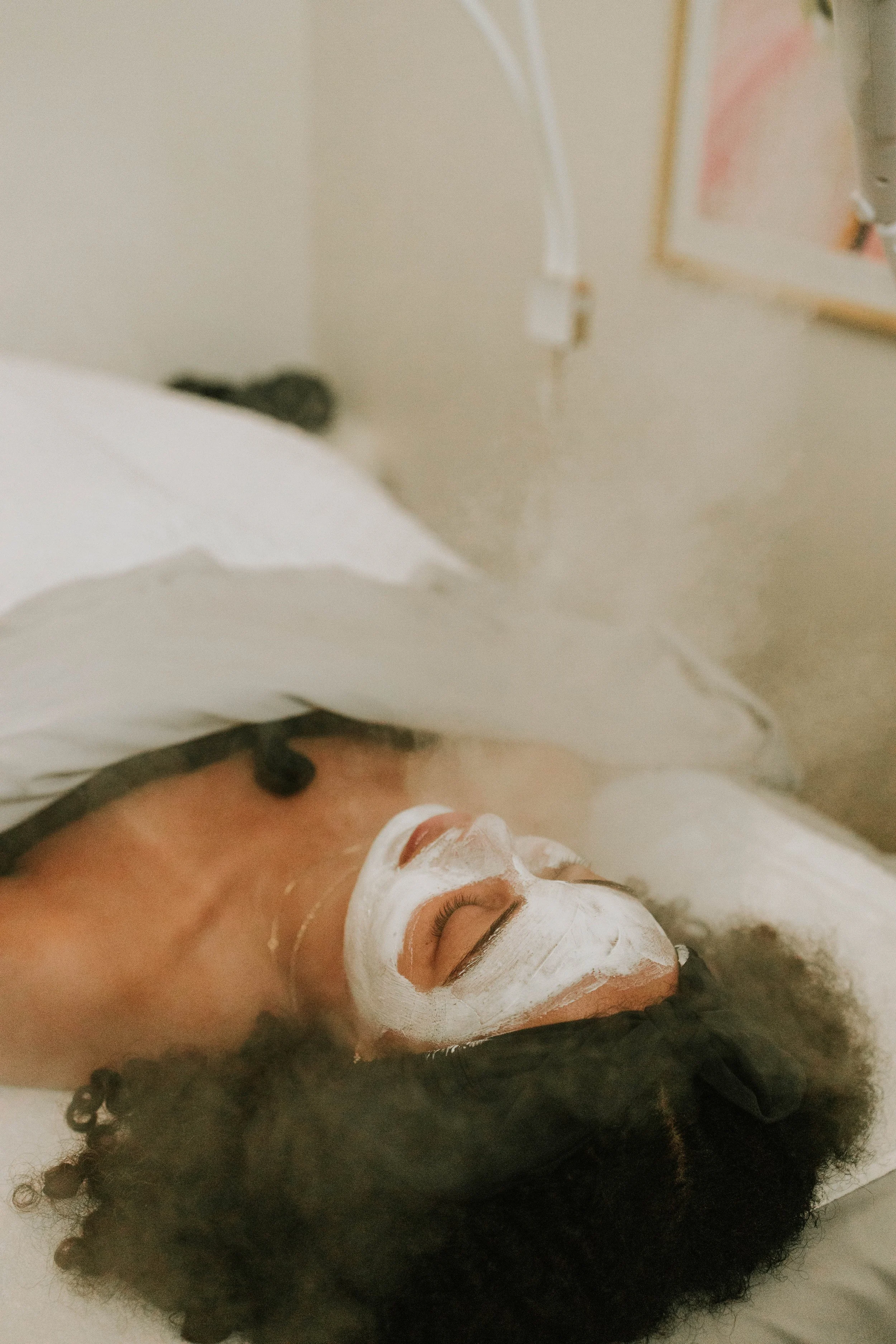 A person lying down with their eyes closed, wearing a white facial mask and headband, appearing to be receiving a spa treatment or facial therapy.